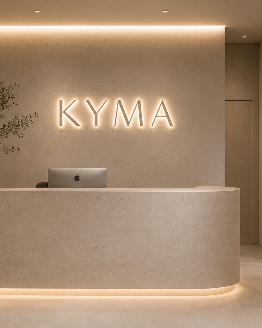 Modern reception area with illuminated 'KYMA' sign on a beige wall behind a curved beige reception desk with an Apple computer, minimal decor, soft lighting