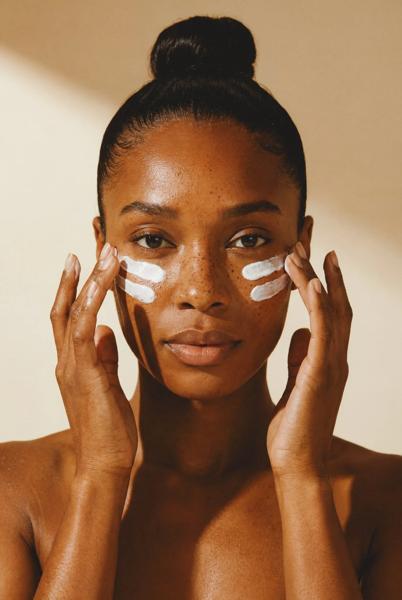 A woman with dark skin and freckles applying skincare cream to her face.