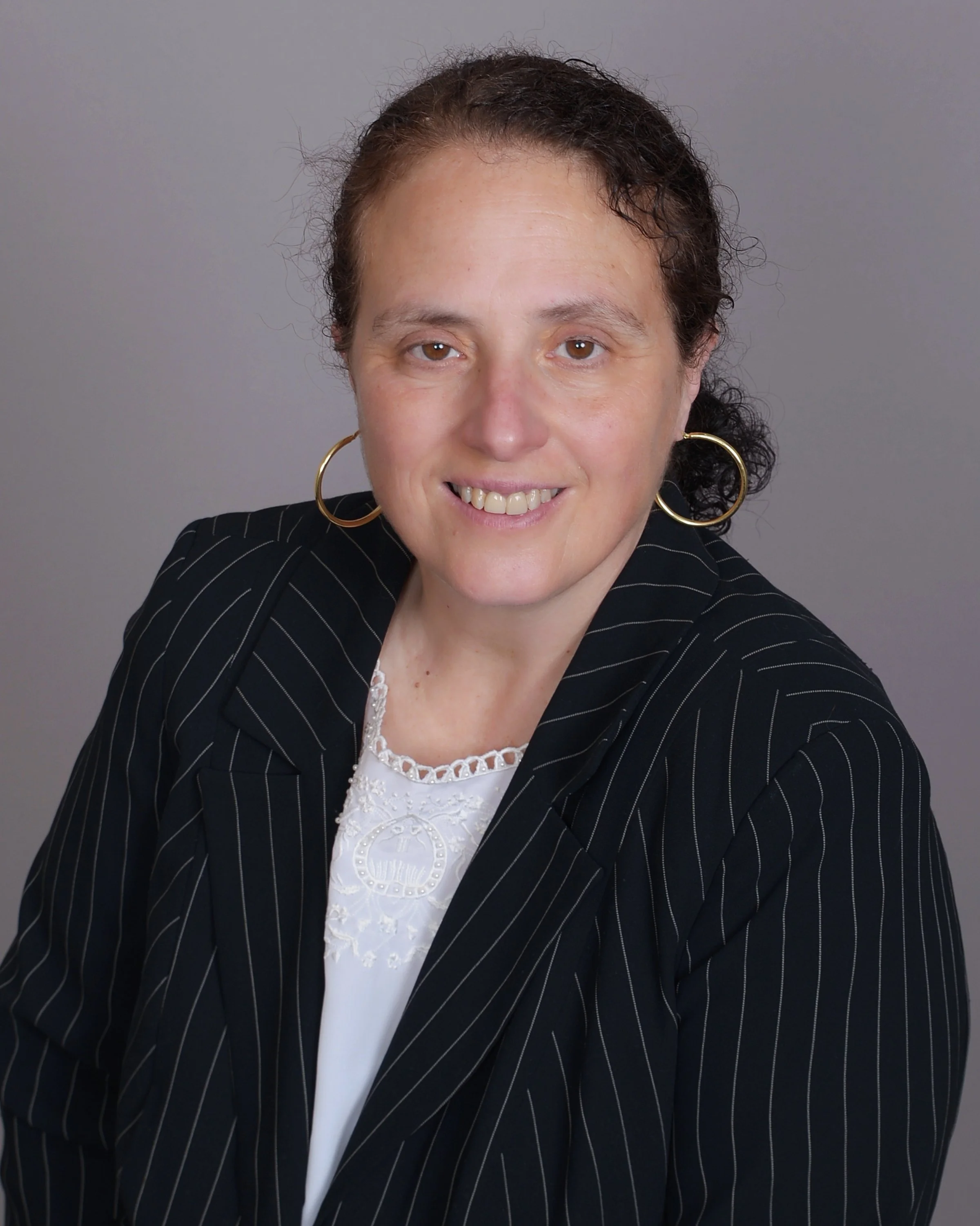 Headshot of a woman with brown hair, wearing large hoop earrings, a black striped blazer, and a white top with lace details, against a gray background.