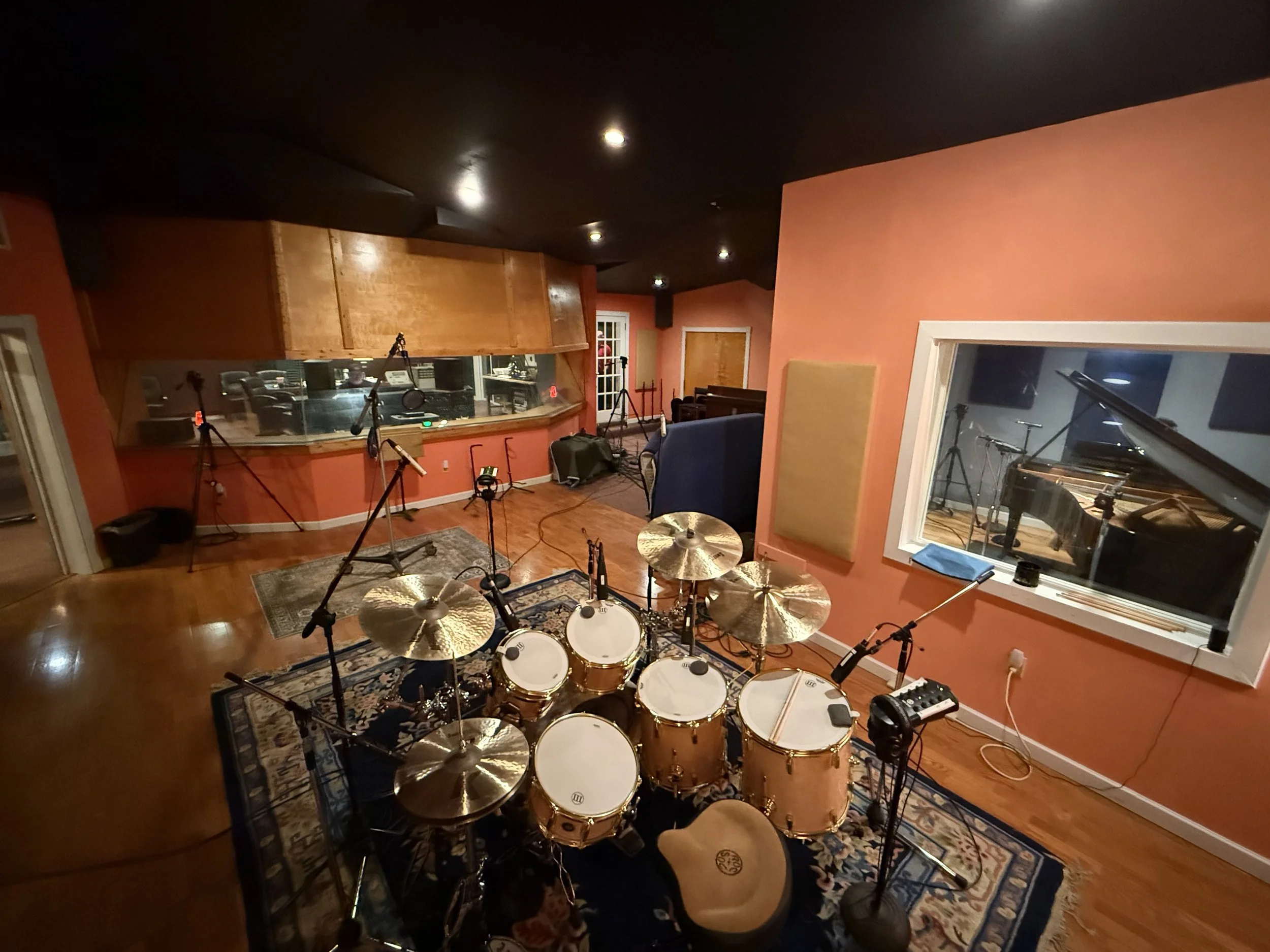 Recording studio with a drums set in the foreground, microphones, and soundproof windows, decorated with orange walls and wooden panels.