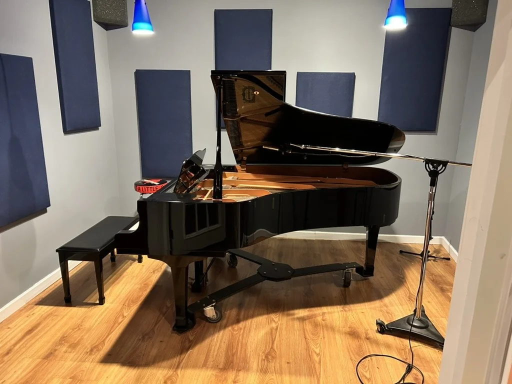 A black grand piano in a music studio with blue acoustic panels on the walls, a black bench, and a microphone stand in front of the piano.