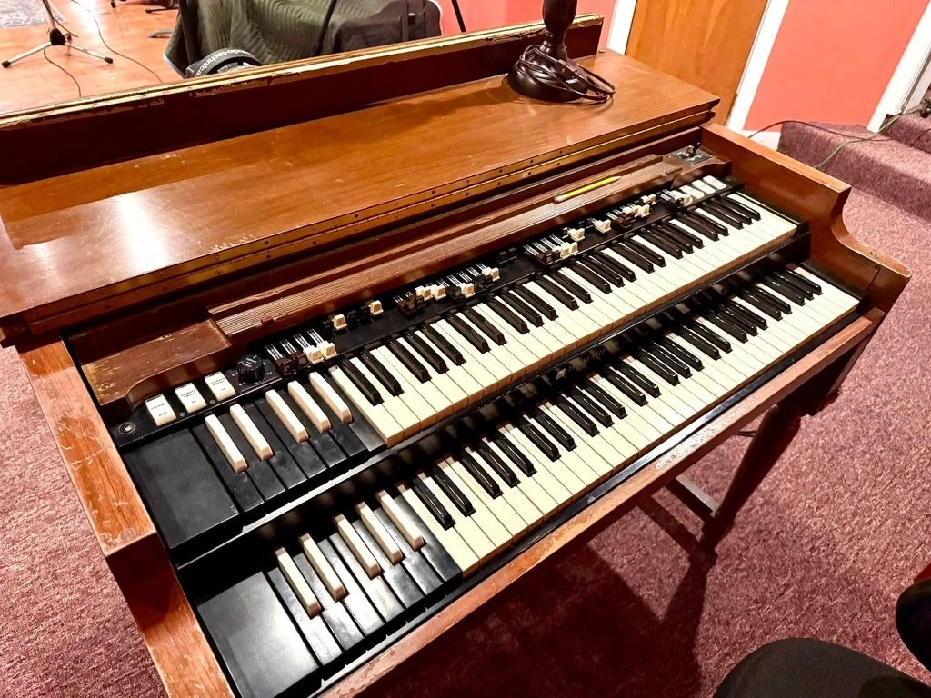 A vintage wooden piano with two rows of keys, featuring various control buttons and switches, situated in a room with pink and orange striped walls and red carpet flooring.