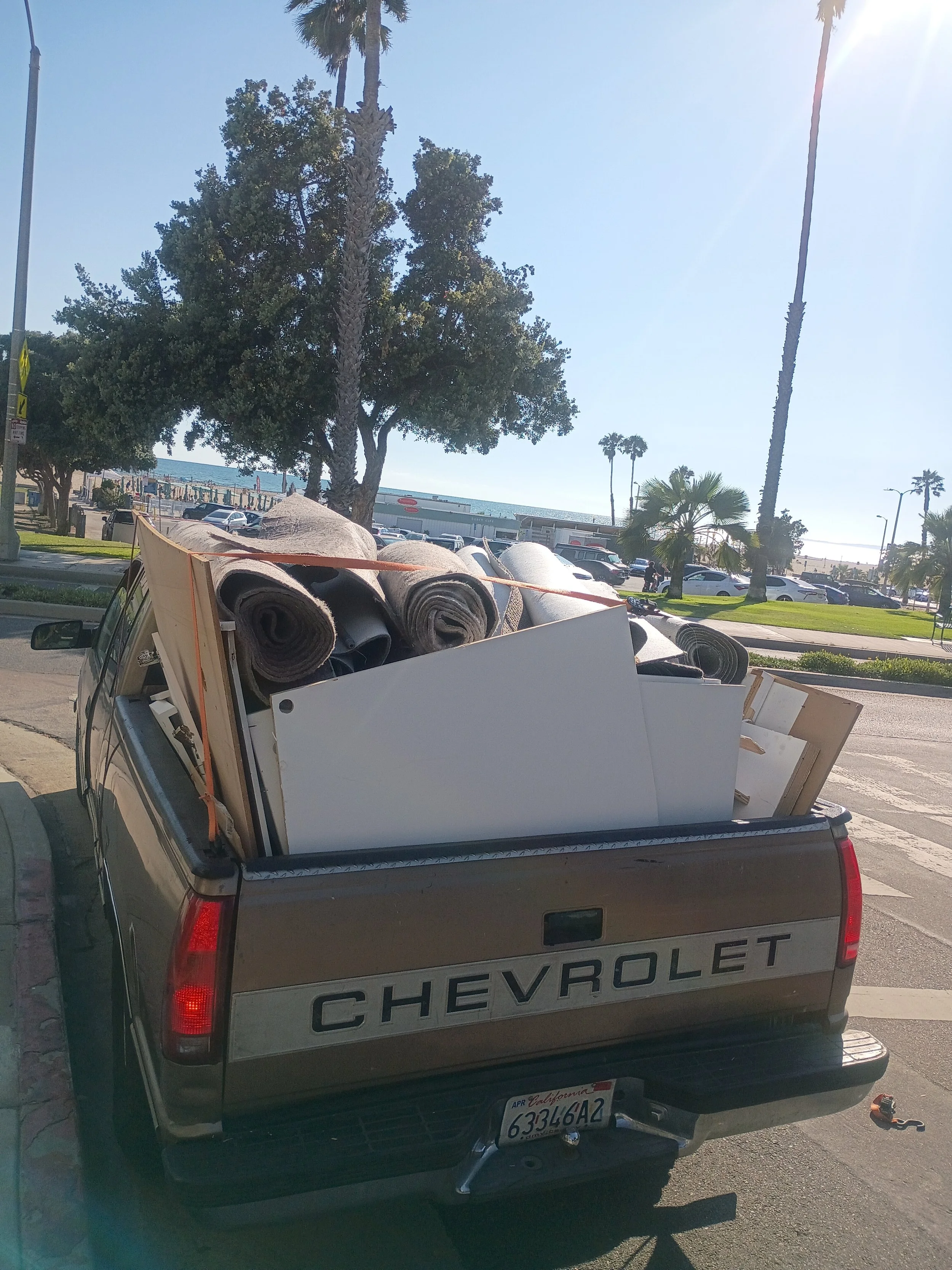 Back of a gray Chevrolet pickup truck loaded with rolled-up carpets, parked on a street with palm trees and a beach in the background.