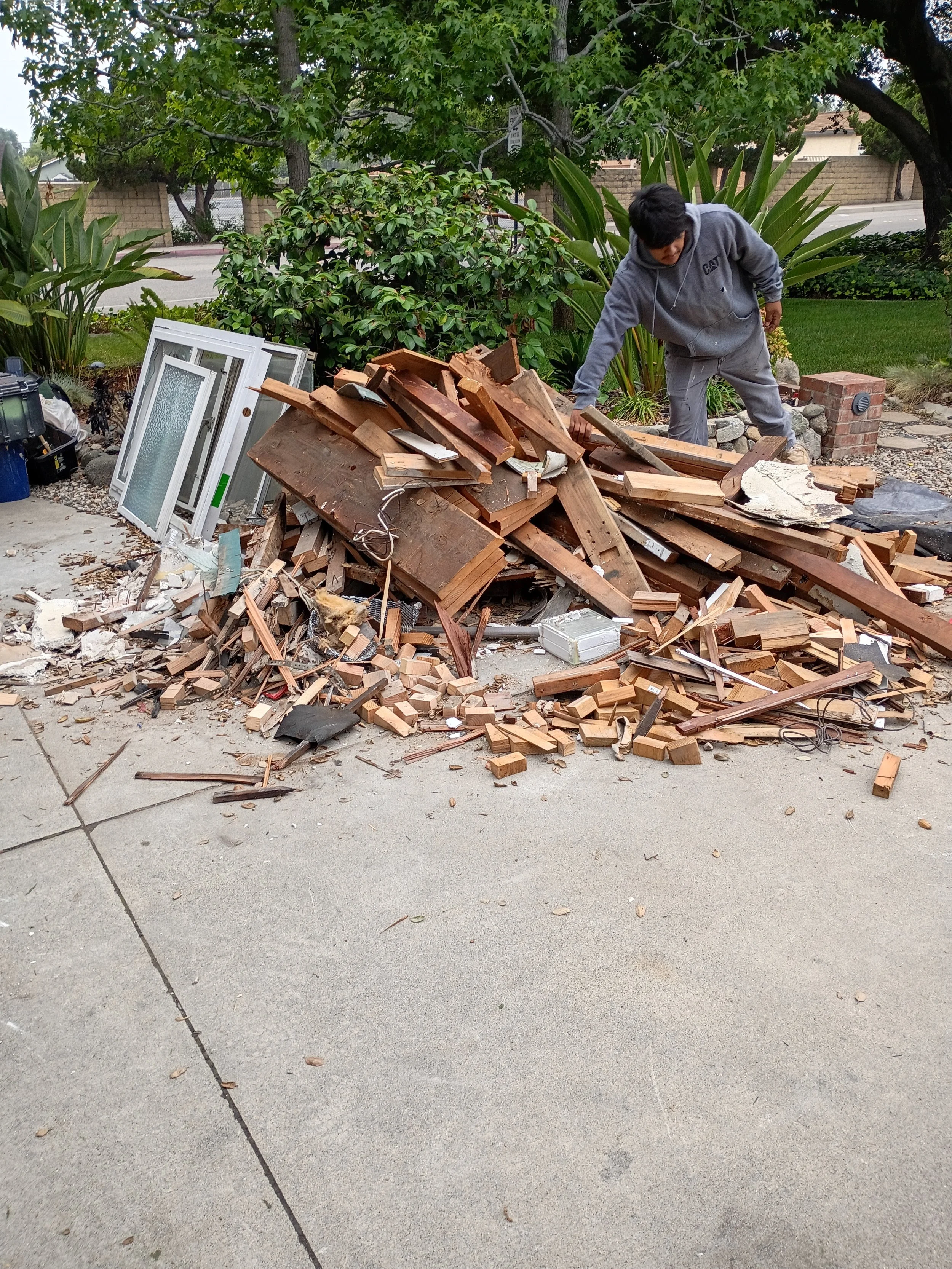 A person wearing a grey hoodie and grey pants is leaning over a large pile of broken wood and debris on a concrete driveway, with a garden and trees in the background.