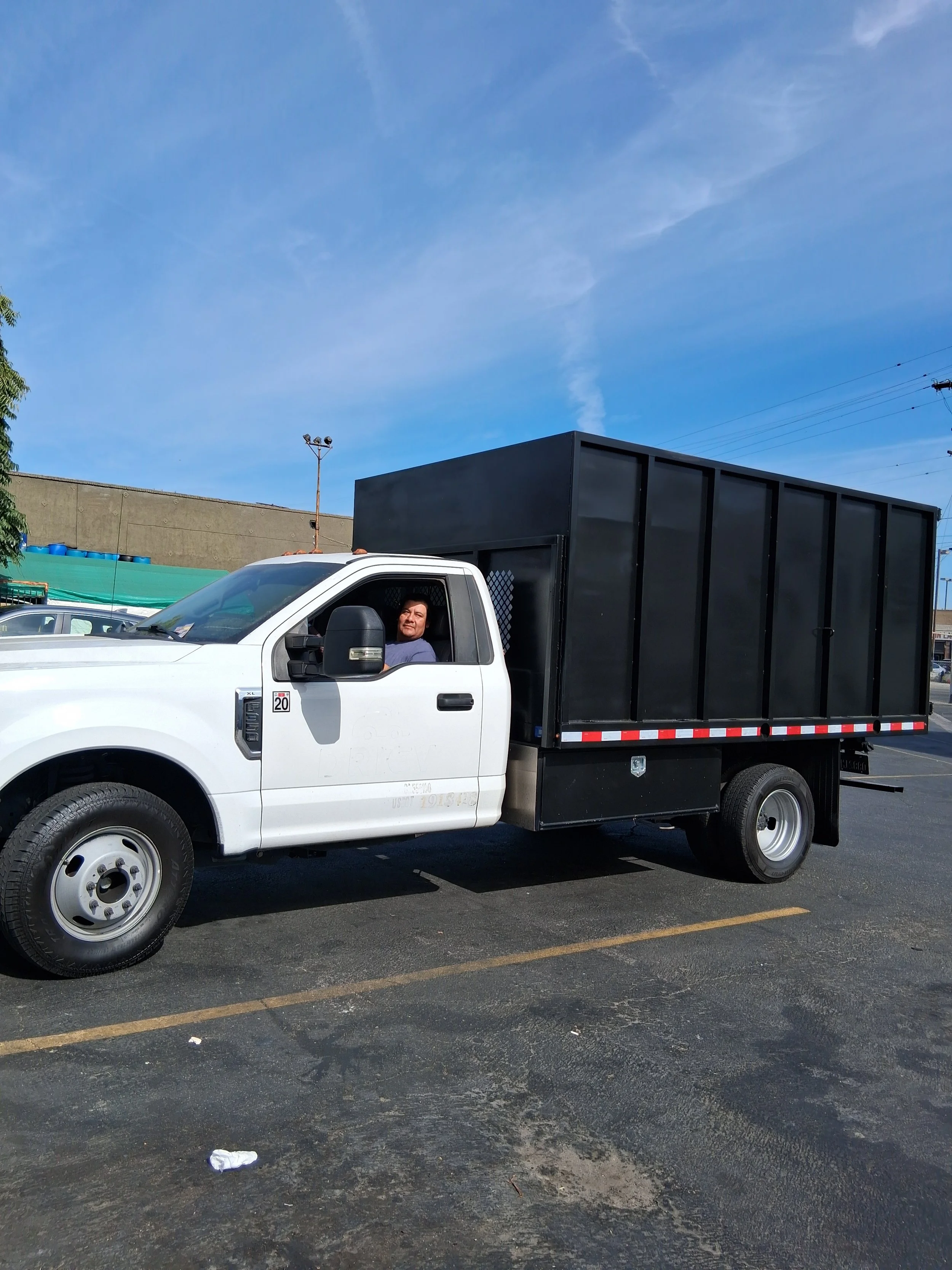 A white pickup truck with a black cargo container on the back, a man in the driver's seat, in a parking lot with a blue sky overhead.