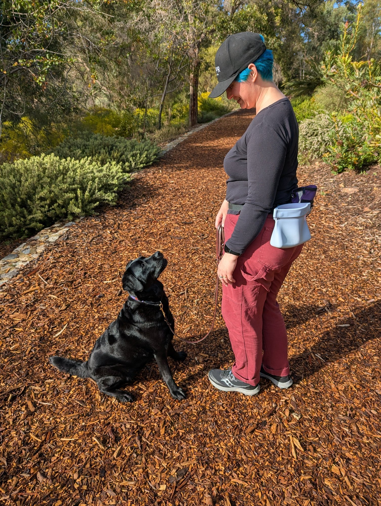 Jasmin smiling down at Lumi, a black Labrador, who sits looking up at her on a bush path