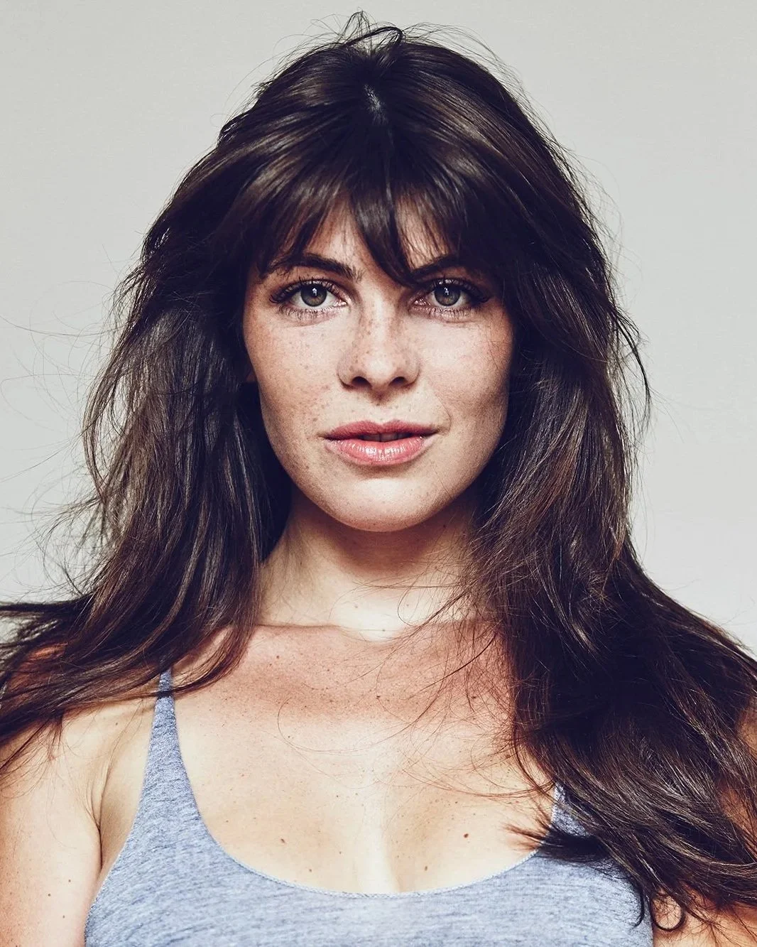 A woman with long, wavy brown hair and blue eyes taking a selfie against a plain white background.