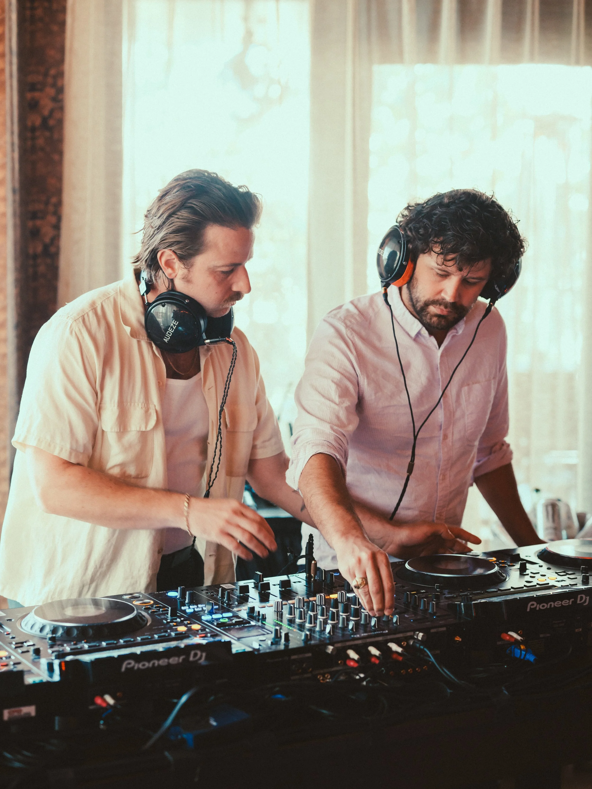 Two men DJing together with DJ equipment, wearing headphones in a room with large windows and curtains.