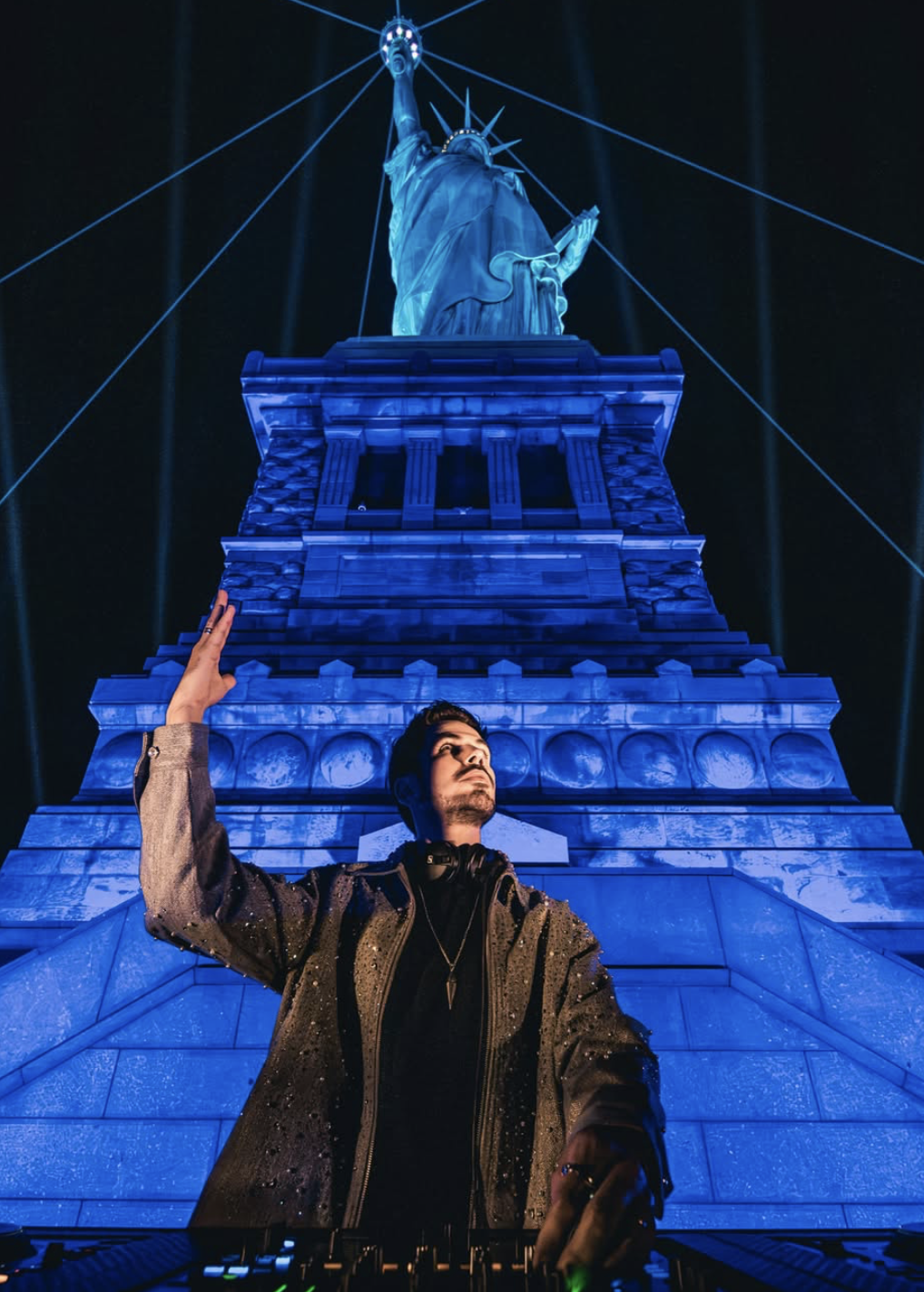 A man in a dark jacket standing in front of a large illuminated replica of the Statue of Liberty at night, with blue lighting and laser beams in the background.