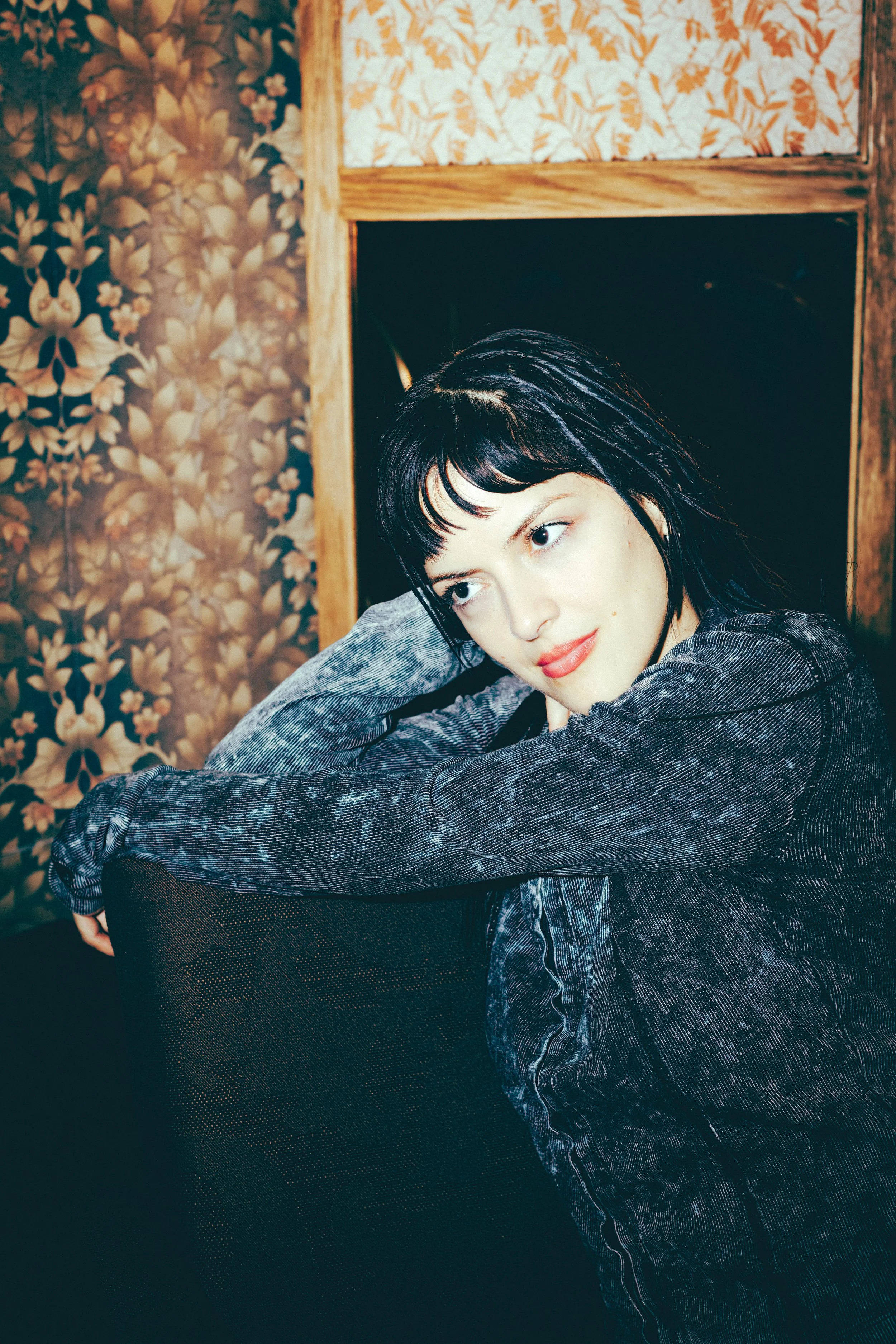 A young woman with black hair and light skin sitting in a dark room with floral and leaf wallpaper, wearing a dark velvety jacket, resting her head on her hand, looking to the side with a slight smile.
