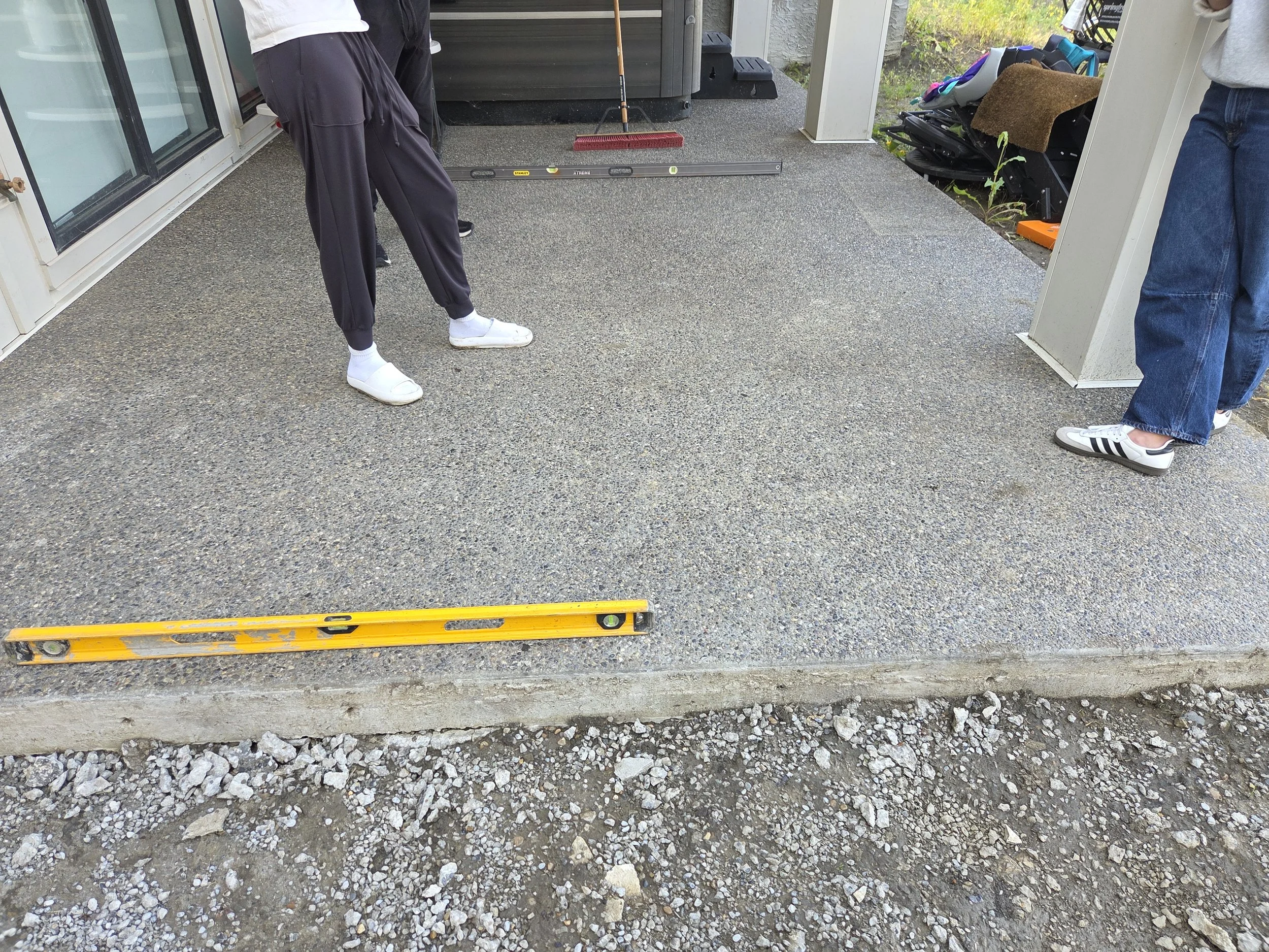 A person in black pants and white shoes standing on a paved surface, near a level instrument on the ground used for construction or surveying, with a yellow rail in front. The background shows a sliding door, some gardening tools, and rocks or gravel
