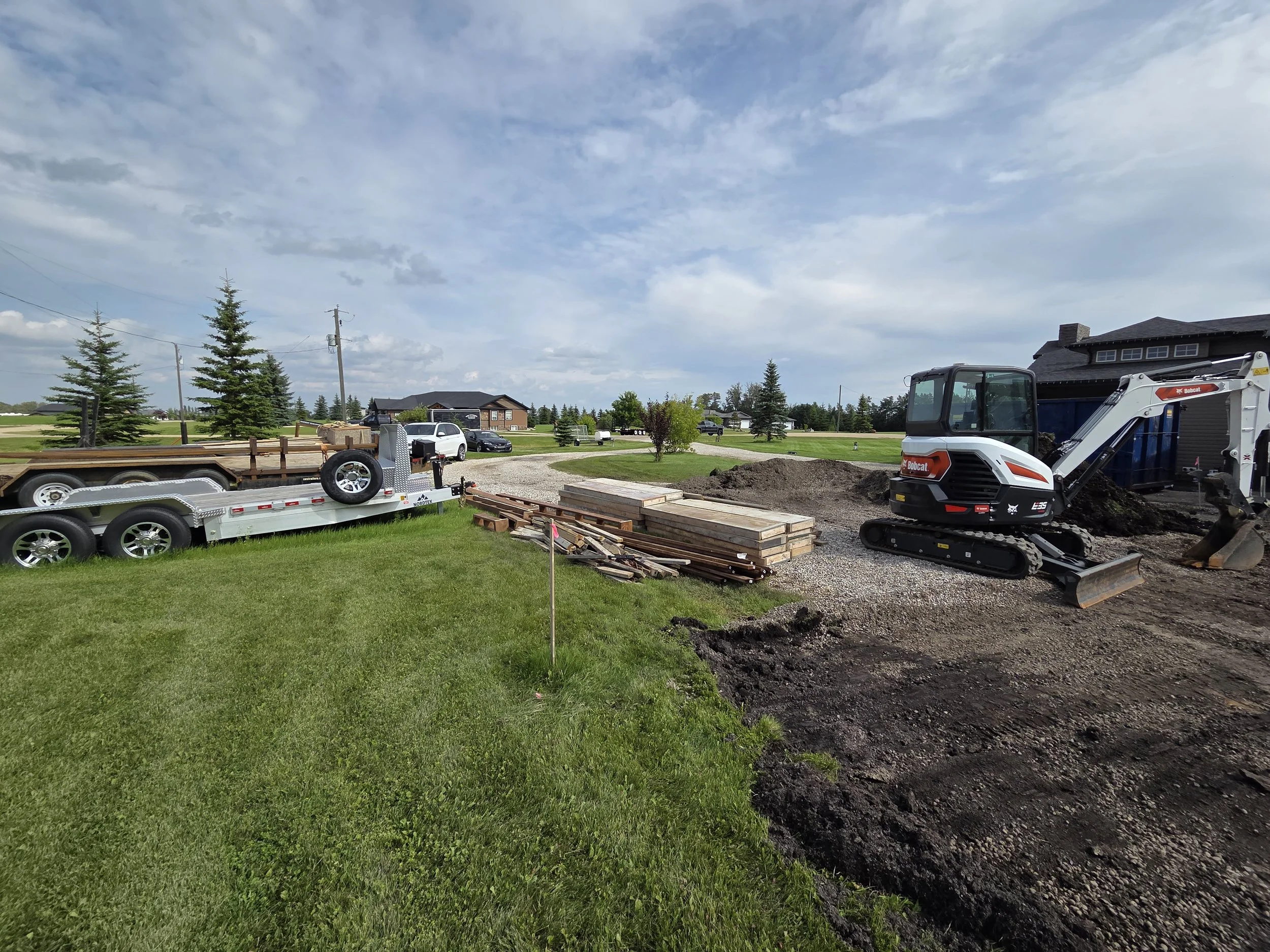 A complete commercial-grade concrete project site mobilization featuring heavy flatdeck trailers, wood formwork materials, and a Bobcat compact tracked excavator in the Parkland County, Spruce Grove area.