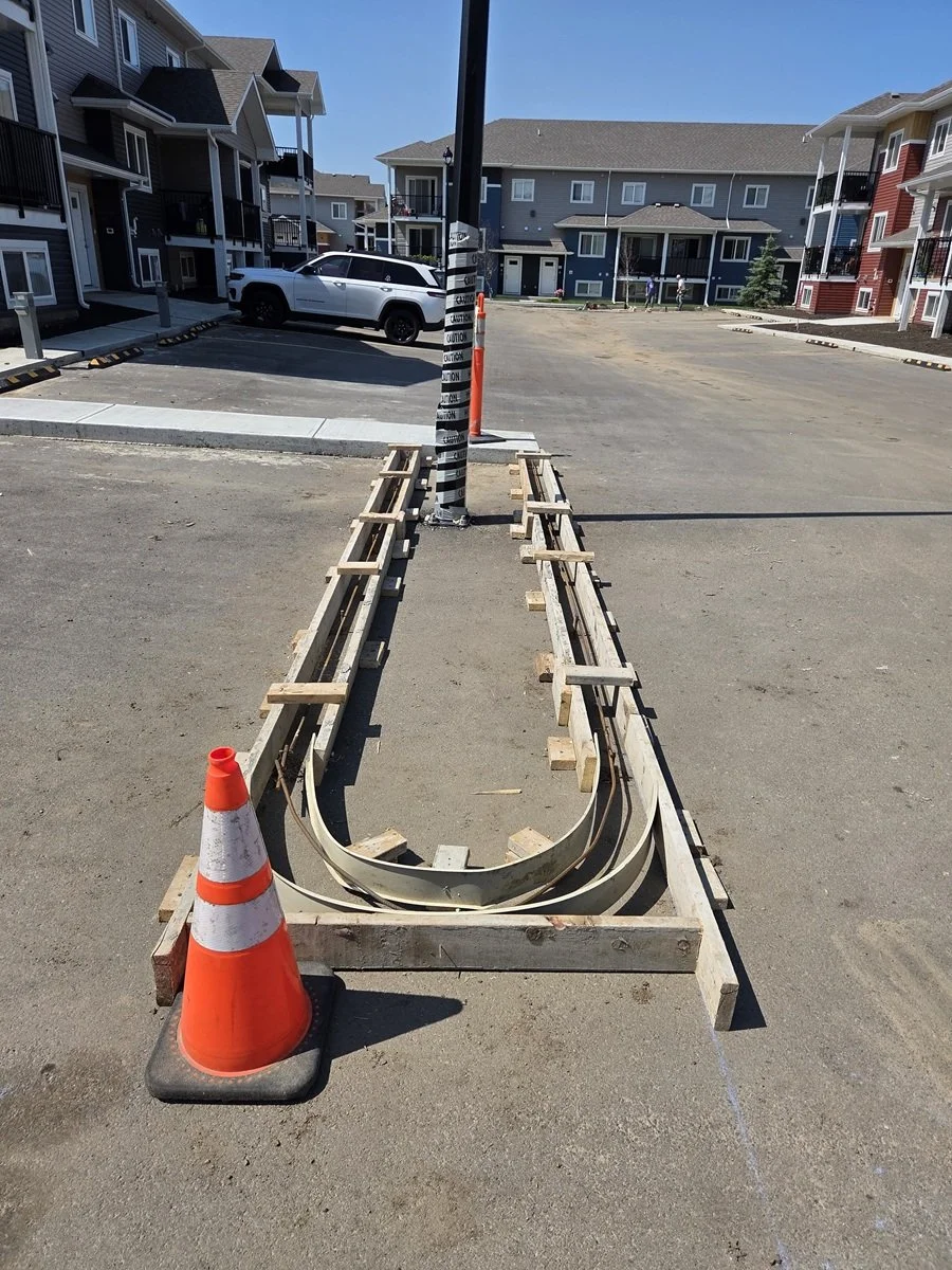 Underside of a train or tram track under construction with wooden forms, orange traffic cone, and caution tape wrapped around a pole in the center. Apartment buildings in the background.