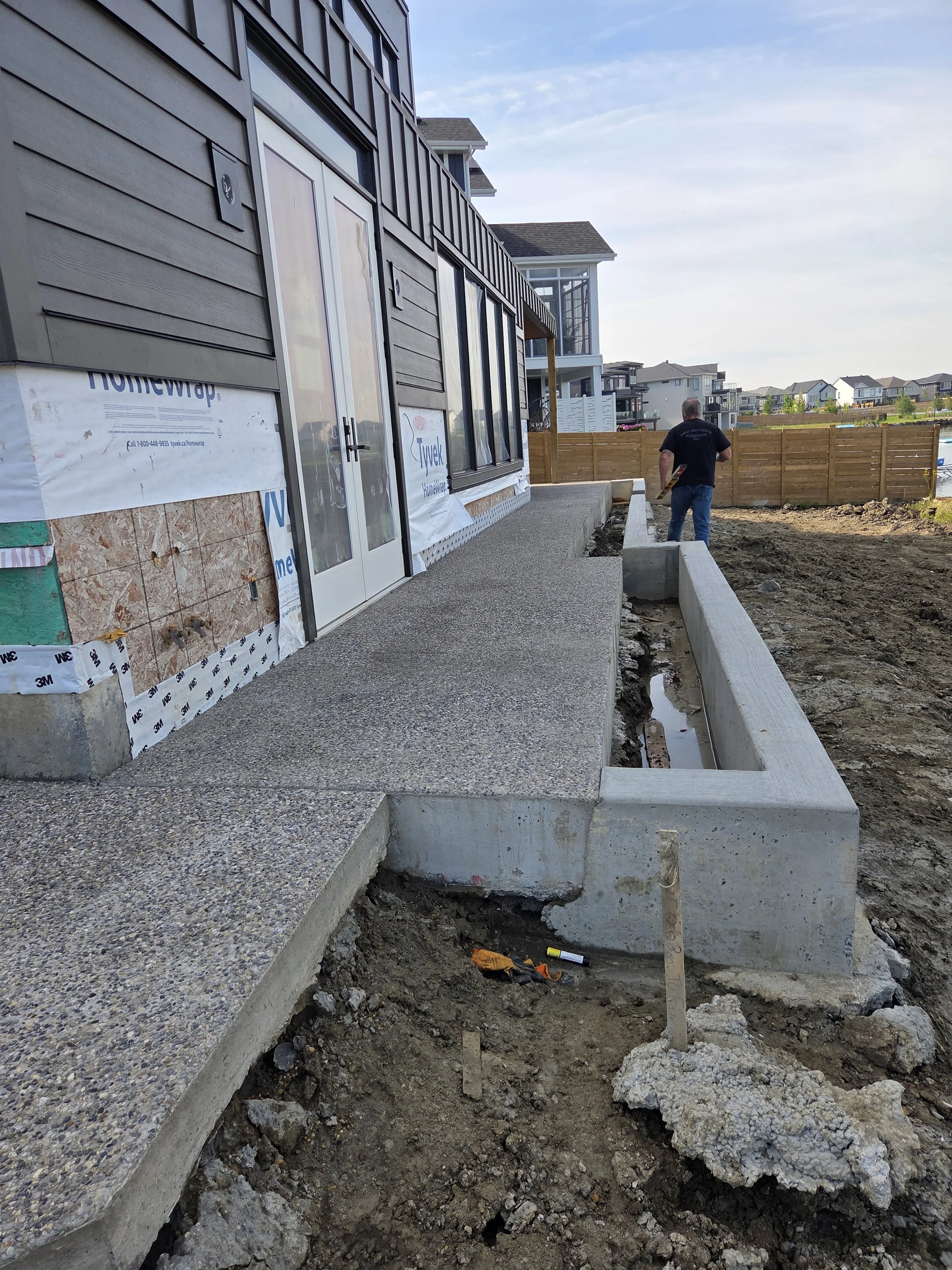 A construction worker walking next to a newly poured concrete walkway at a house under construction, with exposed soil and foundation trench in the foreground.