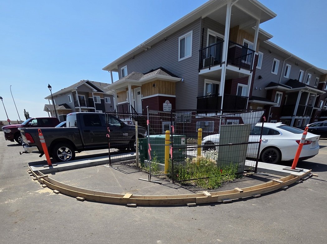 Construction zone in parking lot with temporary wooden curb, metal fence, and construction cones around an area with plants and utility boxes, in front of residential apartment buildings.