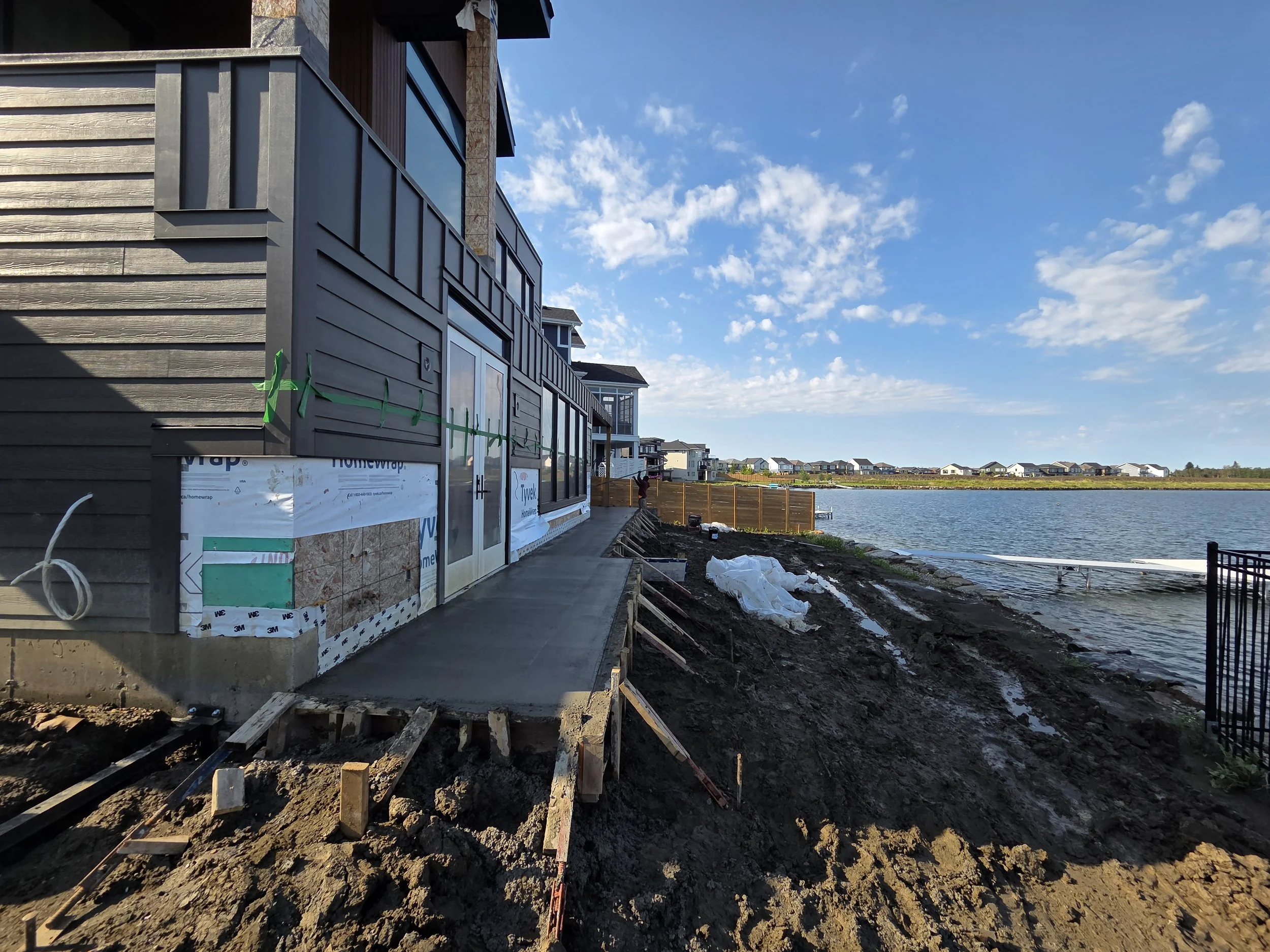 Construction site next to a body of water, with a house under construction featuring large windows and exterior siding, and an unfinished concrete or stone foundation area with wooden formwork, during daytime with blue sky and clouds.