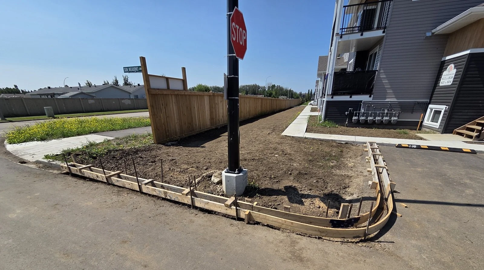 A sidewalk with a curb being constructed around a black pole with a stop sign, next to a residential building with balconies, electrical meters, and a wooden fence on a sunny day.