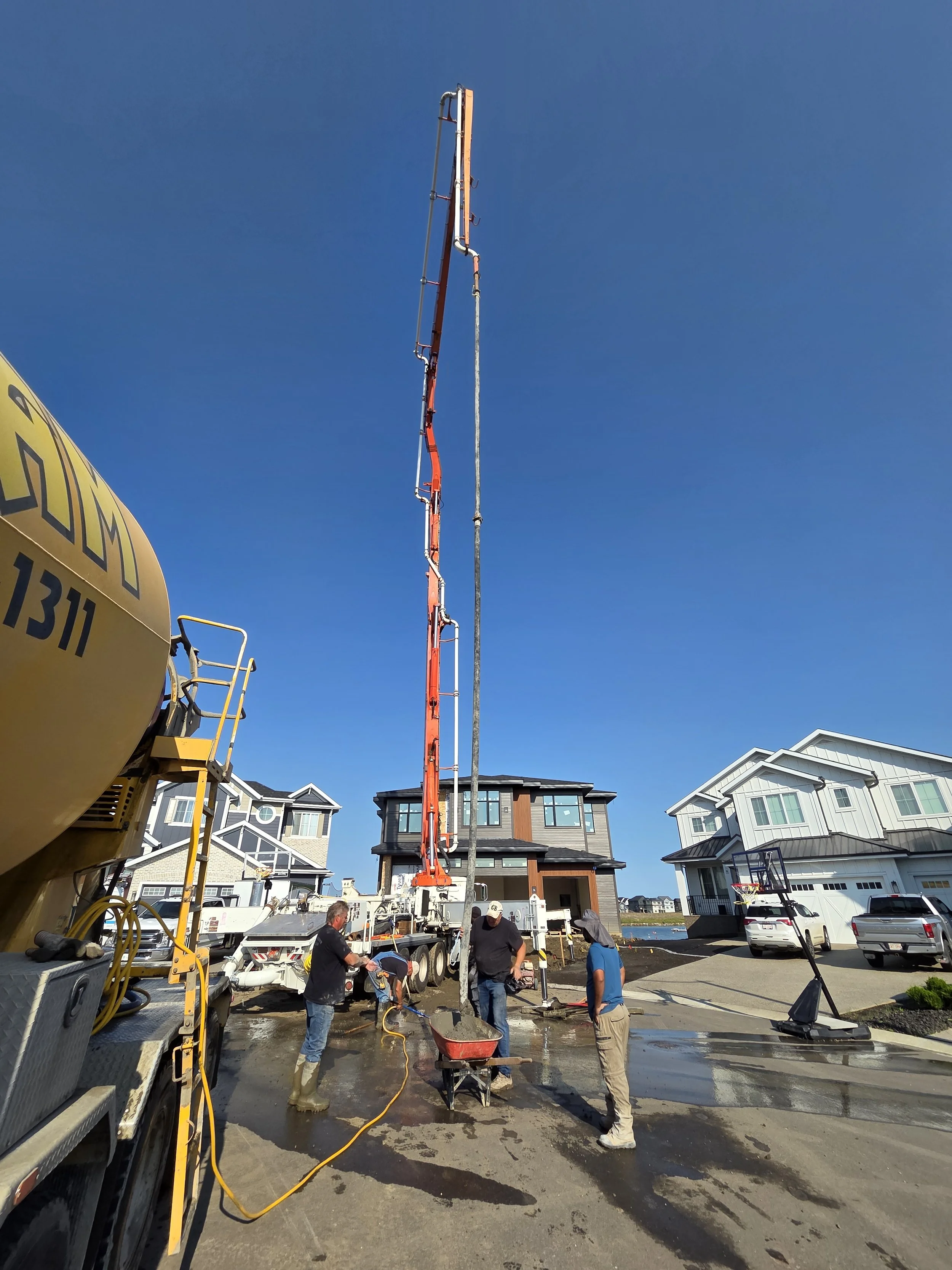Workers are pouring concrete on a residential driveway with a concrete pump truck, a wheelbarrow, and construction materials around, in front of modern houses under a clear blue sky.