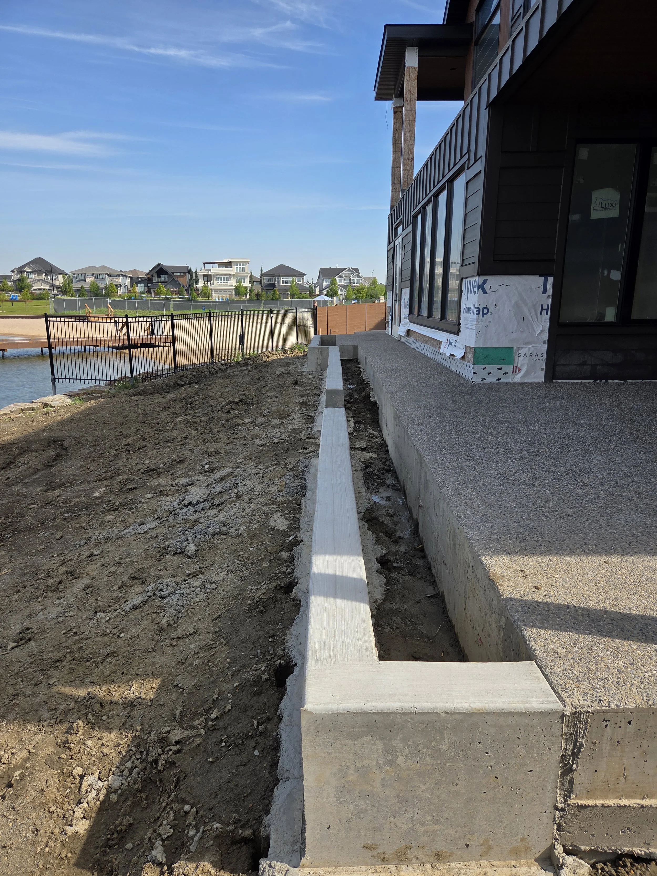 Construction site showing a newly poured concrete walkway beside a modern house with large windows, with a fence and neighboring houses in the background.