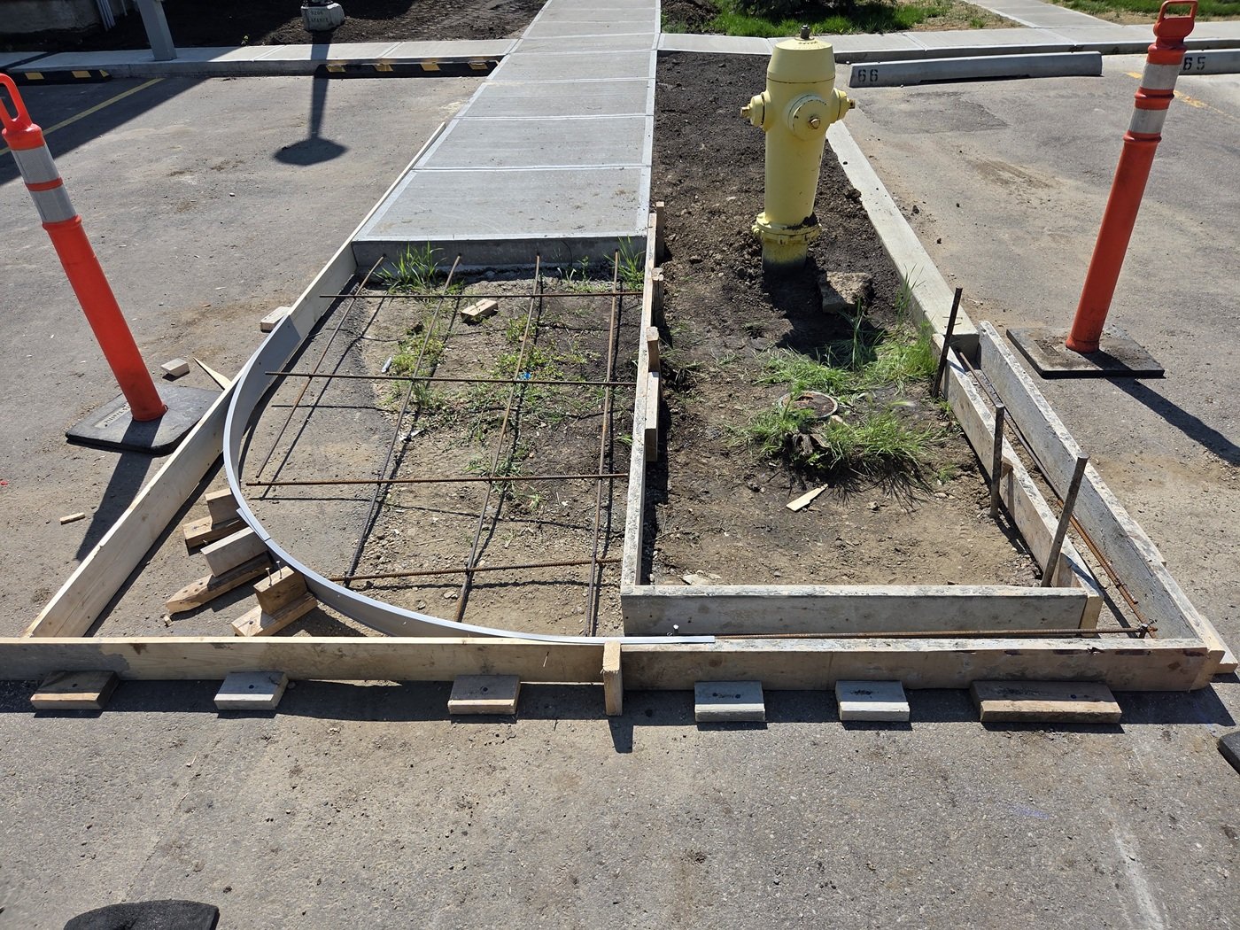 Construction site showing a partially built sidewalk with concrete pavers, orange safety cones, a yellow fire hydrant, and a wooden frame around a planting area with some grass and debris.