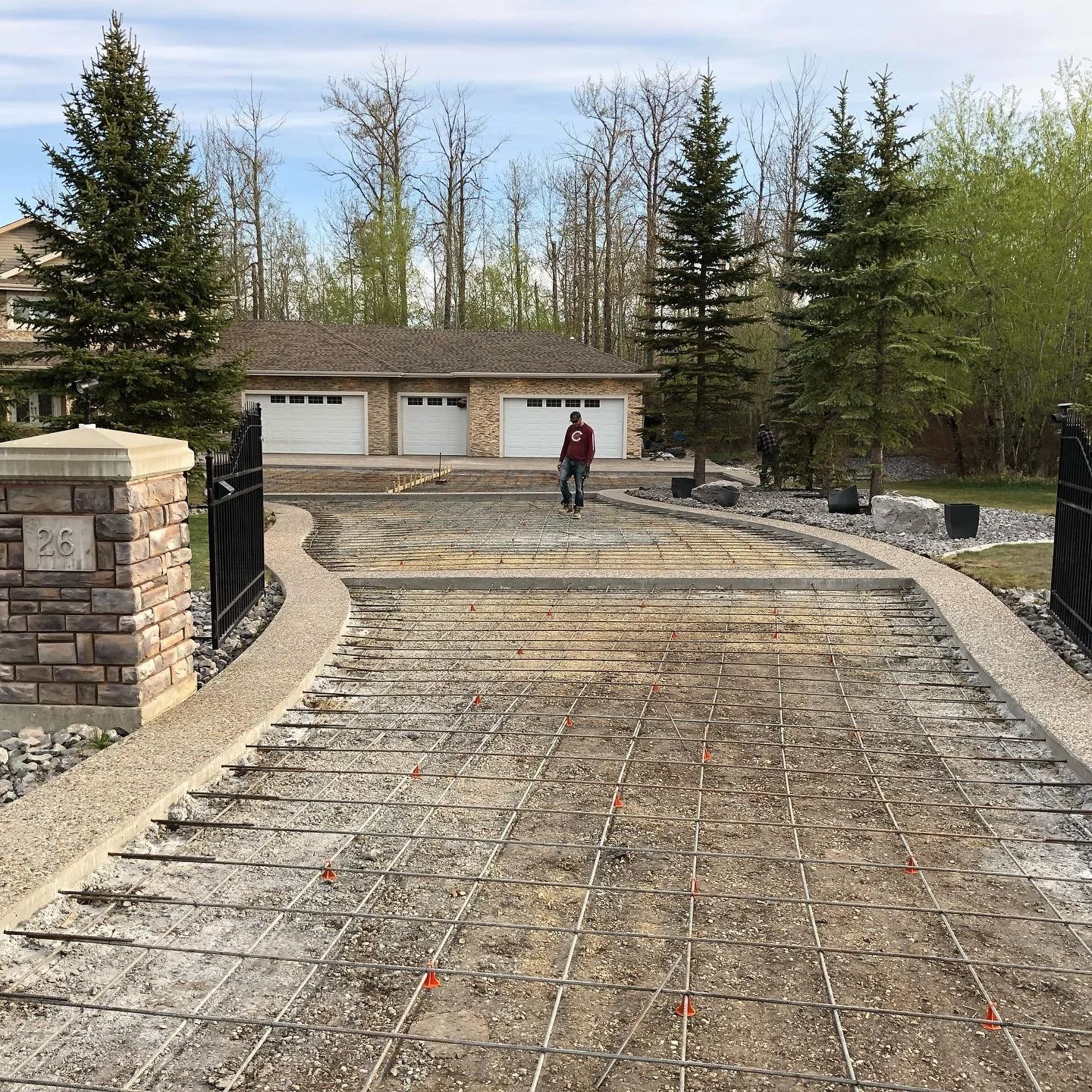 A driveway under construction with a grid of metal rebar laid out, leading to a house with a three-car garage, surrounded by trees and a stone pillar with the house number 26.