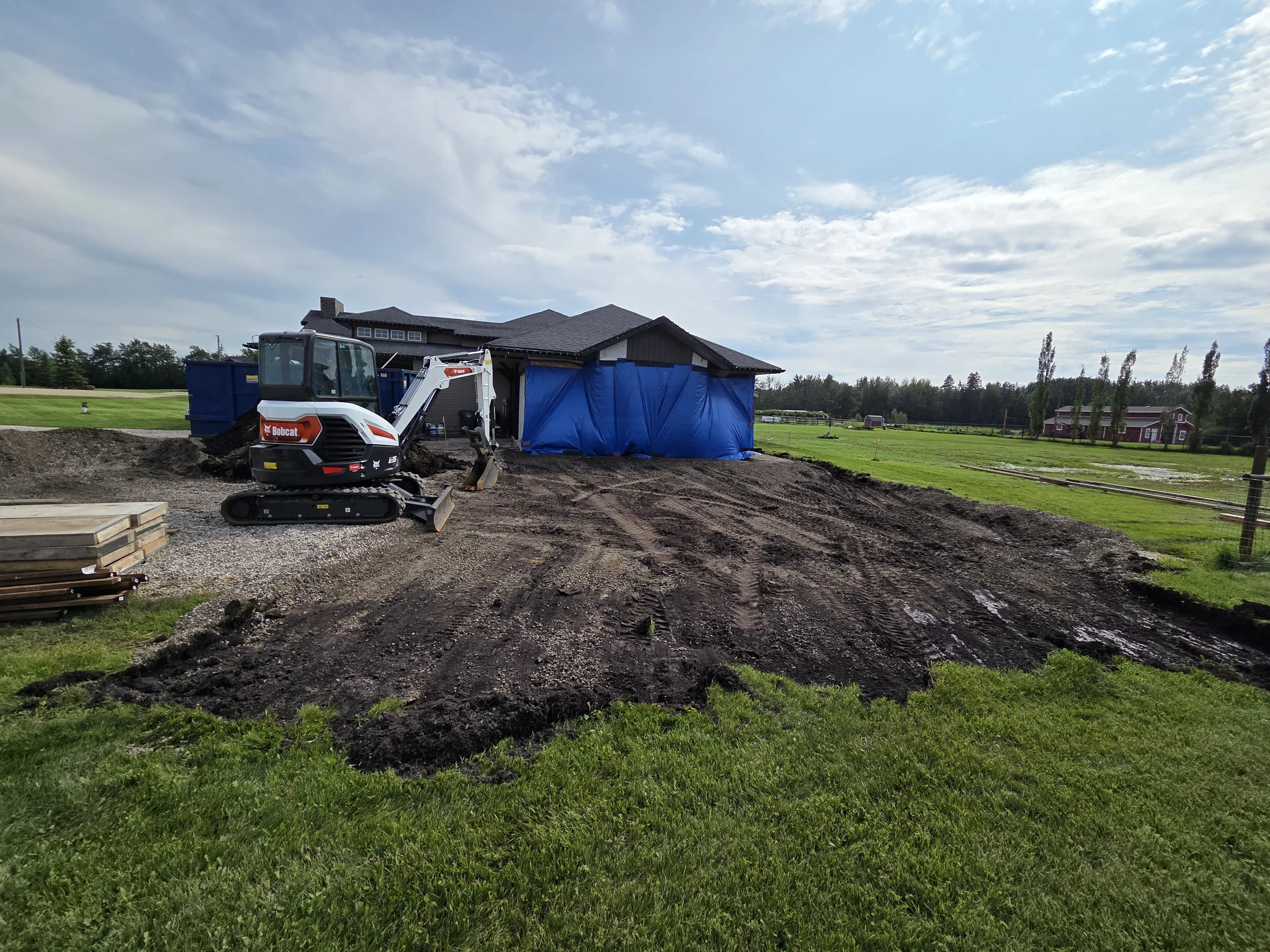 Construction site with dirt and gravel, a Bobcat compact excavator, and a house with a blue tarp covering part of it, surrounded by grass and fields under a partly cloudy sky.