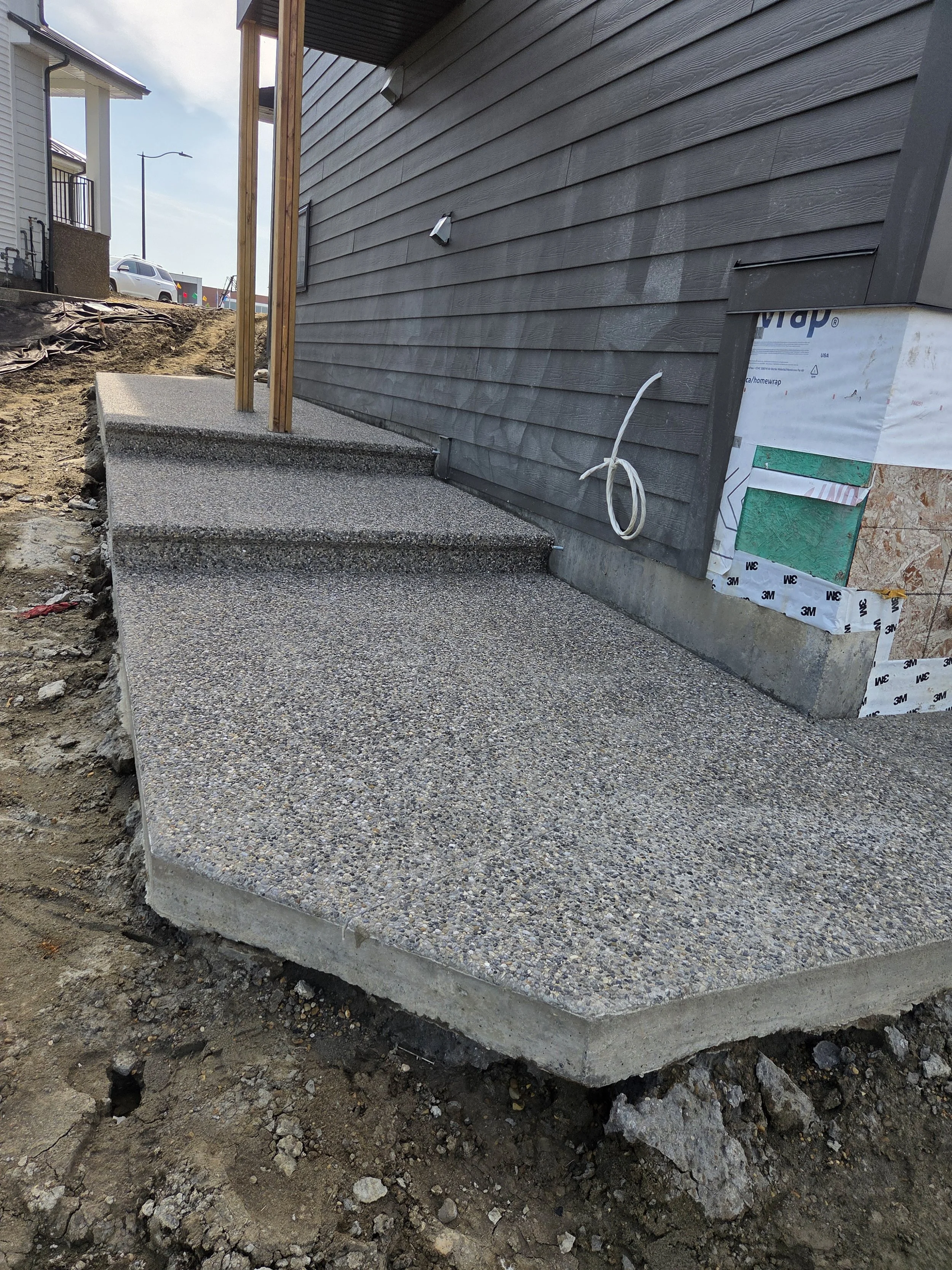 Concrete stairs leading up to a house with a gray wooden exterior. Construction is ongoing, with exposed dirt, rocks, and building materials visible around the stairs. A wooden post supports an overhang at the top of the stairs, and part of a neighbo