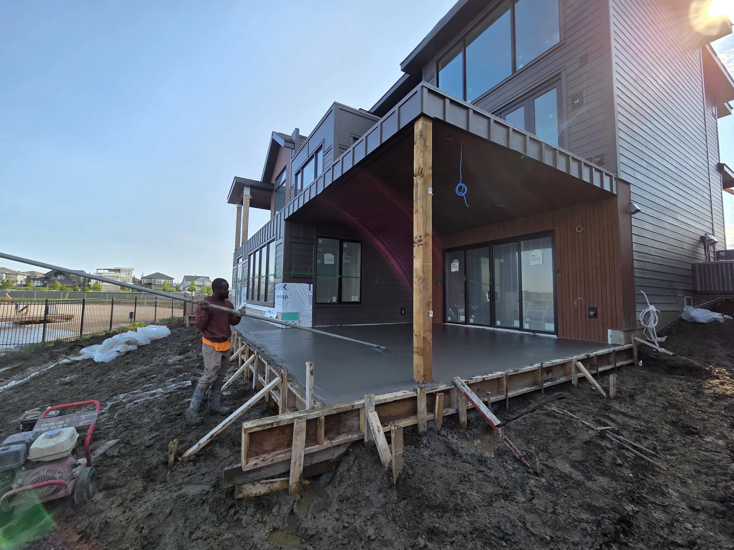 Construction site of a modern multi-story house with a concrete patio and unfinished wooden framework, cloudy sky, and a person working with construction tools.
