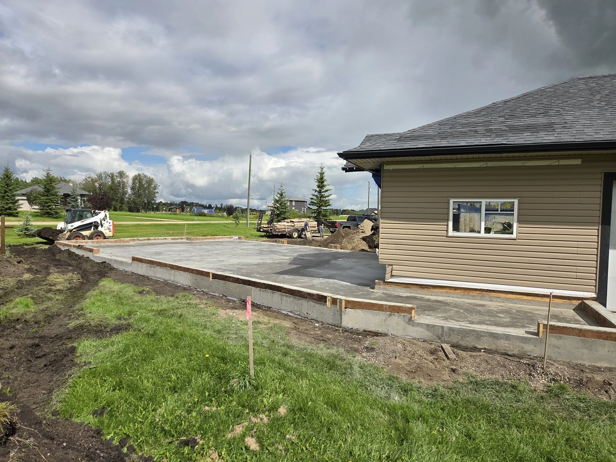 A professional concrete contractor crew using a skid steer for final grading and site cleanup around a massive newly poured residential driveway slab on a Parkland County acreage near Spruce Grove.