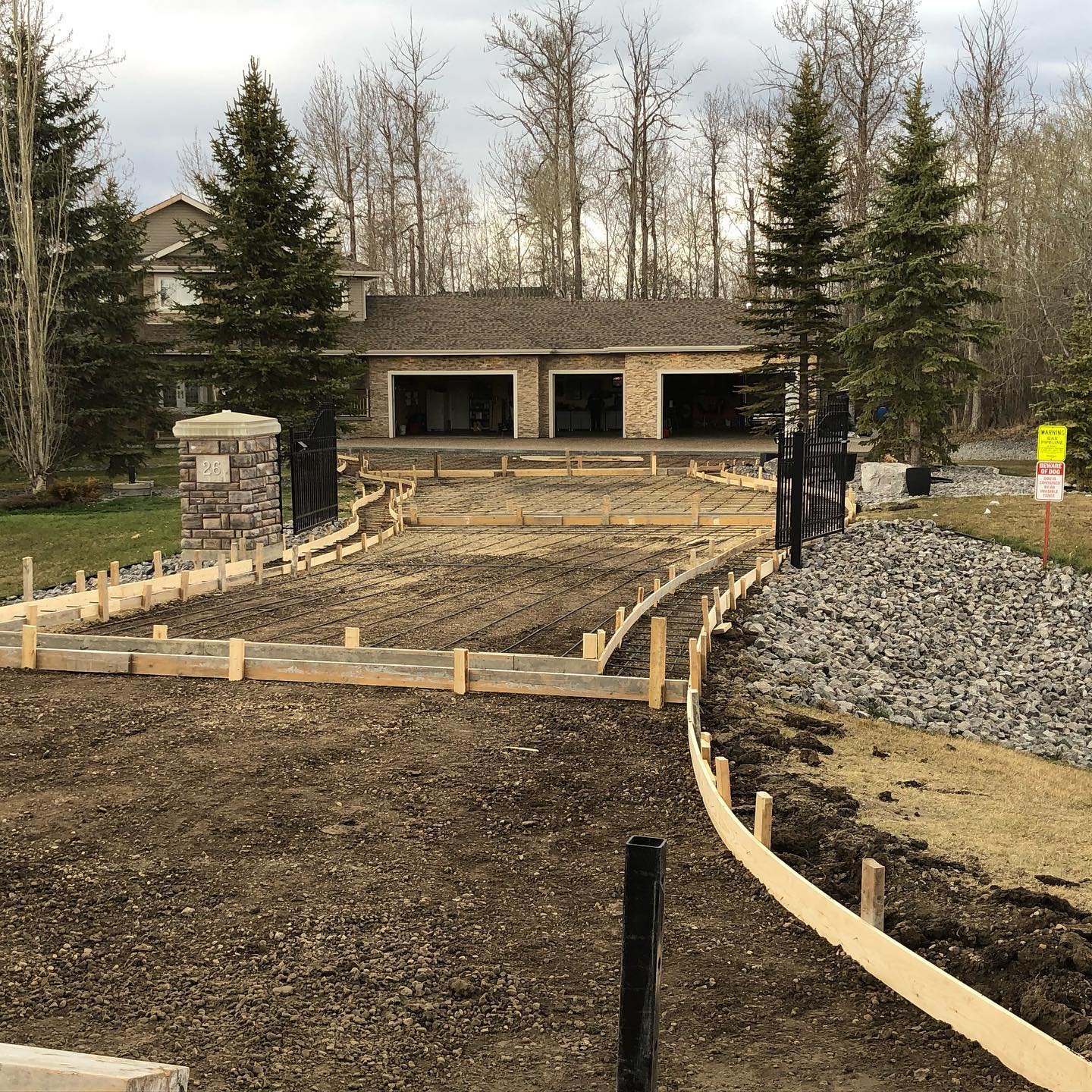 Residential driveway under construction with wooden framing and gravel, leading to a house with trees in the background.