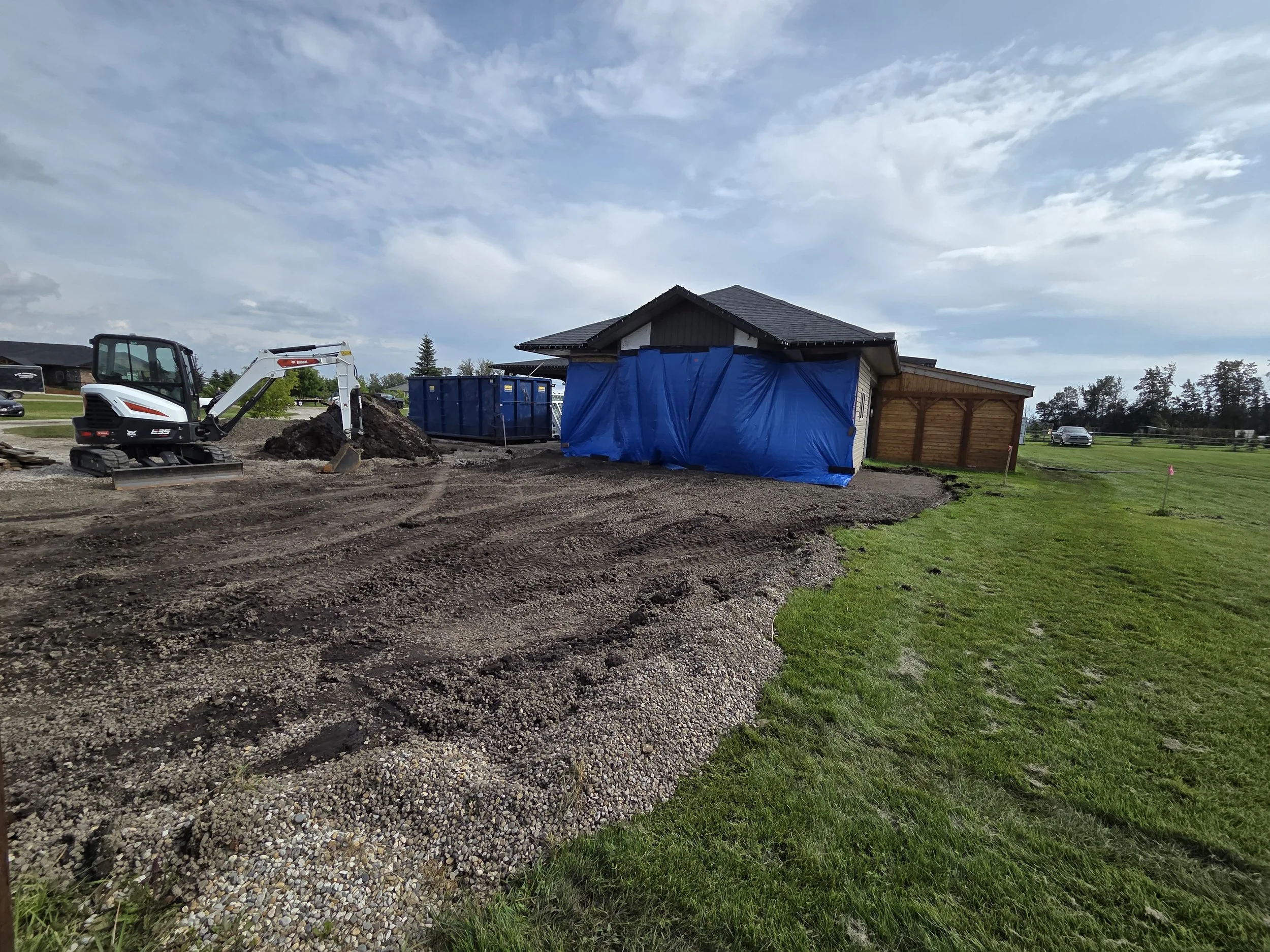 Construction site showing a small excavator digging into the dirt near a house, with a blue tarp covering part of the house, and a gravel pathway in the foreground with grass on the sides.
