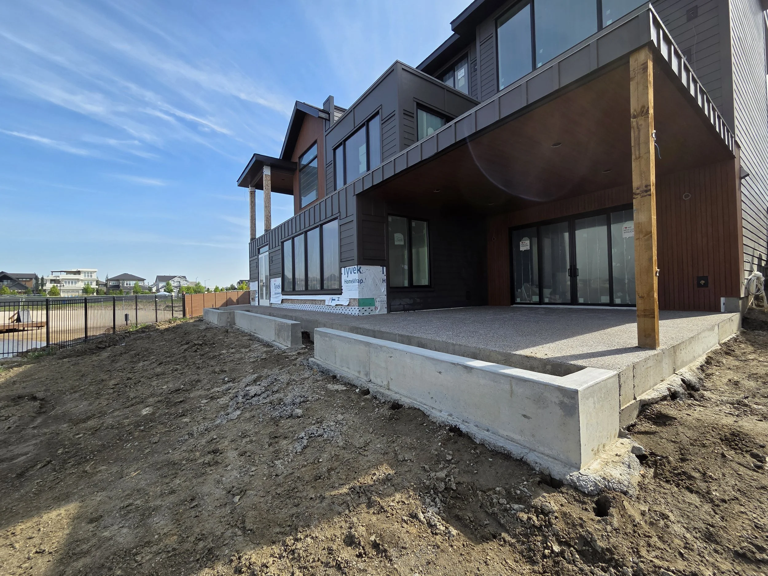 A modern multi-story house under construction with a wooden and metal exterior, large glass doors and windows, and a concrete patio. Construction materials and tools are visible, along with surrounding neighborhood houses.