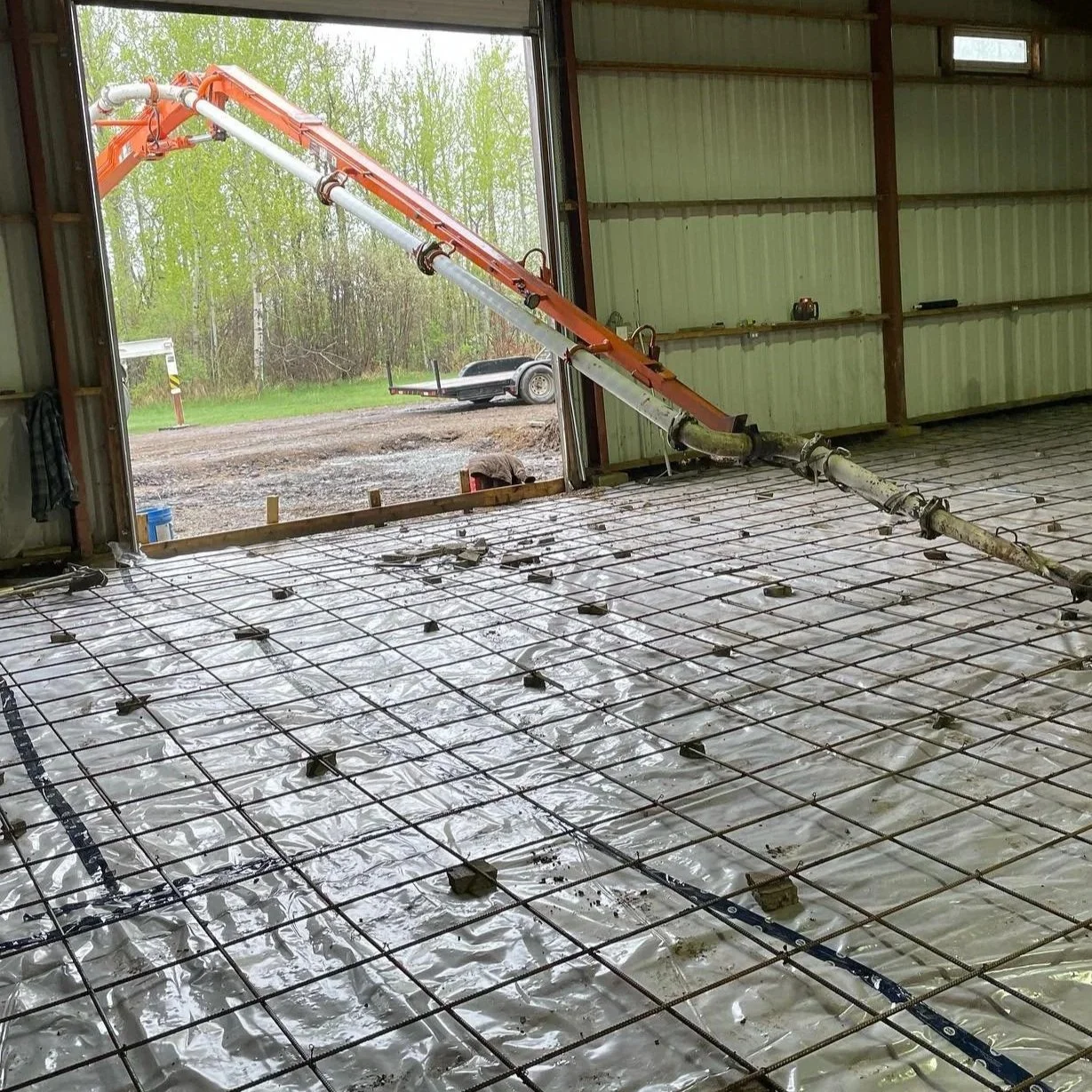 An interior industrial shop floor placement under development showing a concrete boom pump line entering a large bay door over an engineered steel mesh reinforcement grid and vapor barrier in Parkland County.