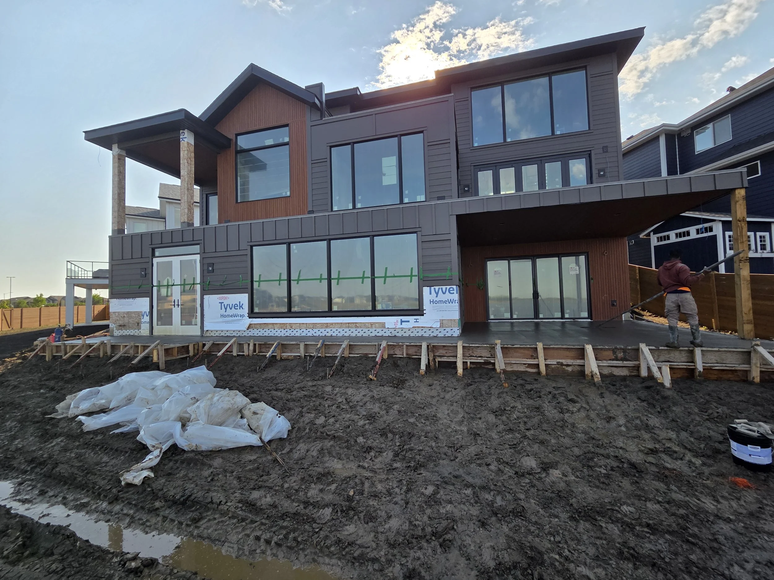 A multi-story house under construction with ongoing exterior work visible. The house features large windows and modern architectural design. Construction worker on site, with some construction materials and tools around, and an unfinished yard with e