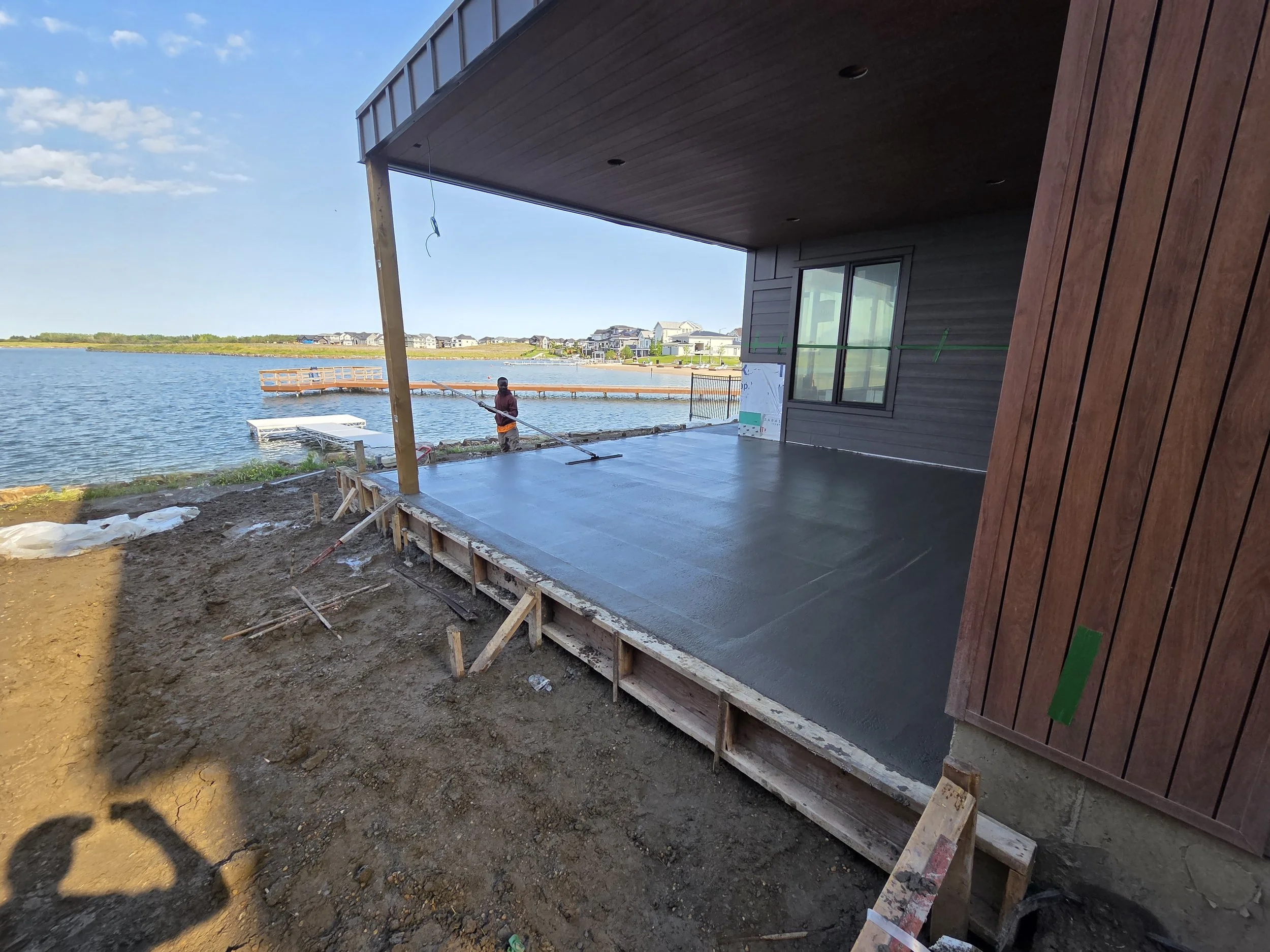 A worker leveling freshly poured concrete on a patio or deck overlooking a body of water, with residential houses in the background at a sunny day.