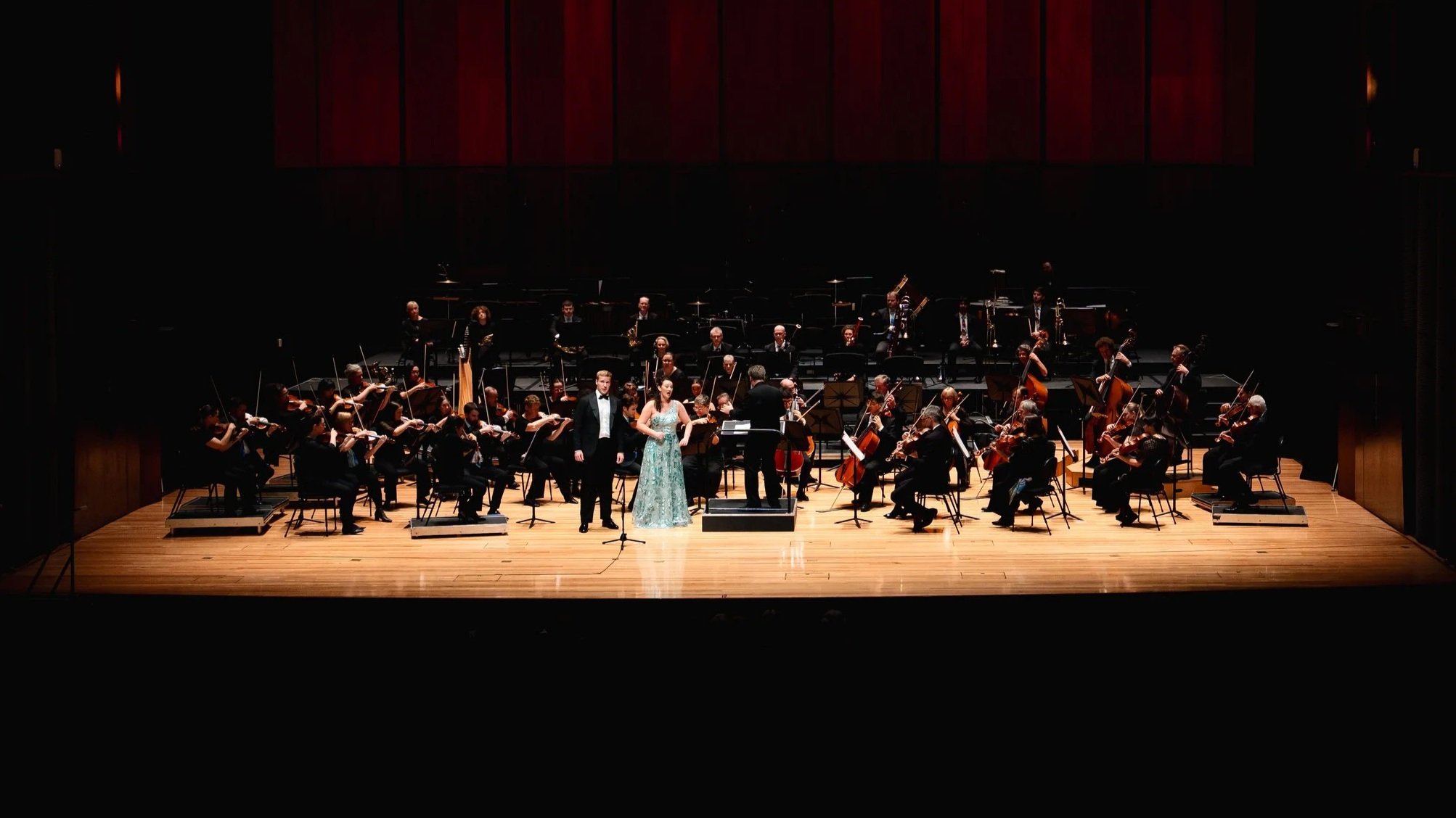 Orchestra performing on stage with two soloists in formal attire, a man and a woman, during a concert in a theater.