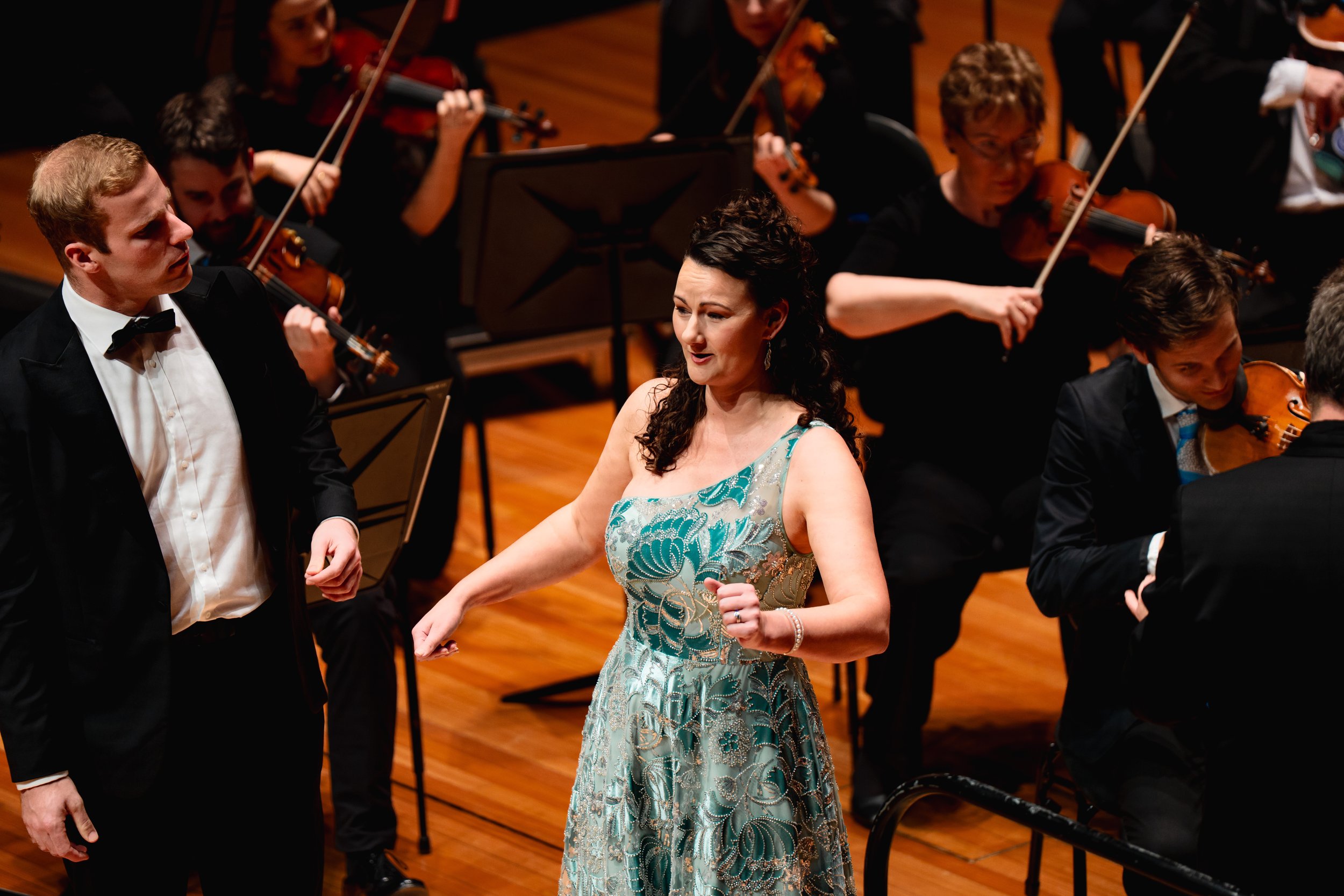 An orchestra performing on stage with a conductor and a female soloist in the foreground wearing a turquoise and gold dress. The orchestra members are playing violins and are dressed in formal black attire.