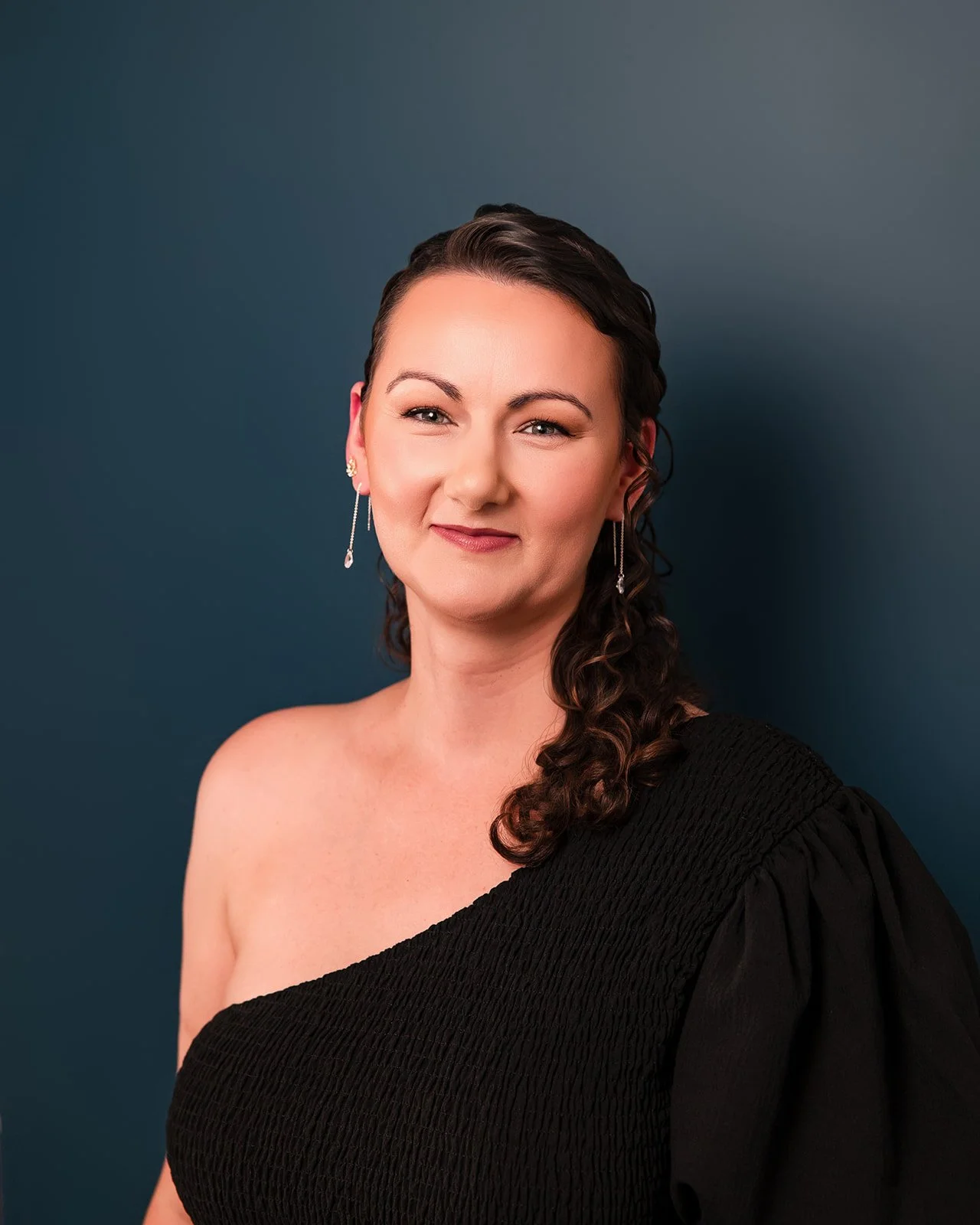 Portrait of a woman with dark, curly hair, wearing a black off-the-shoulder dress and long drop earrings, standing against a dark blue background with a slight smile.