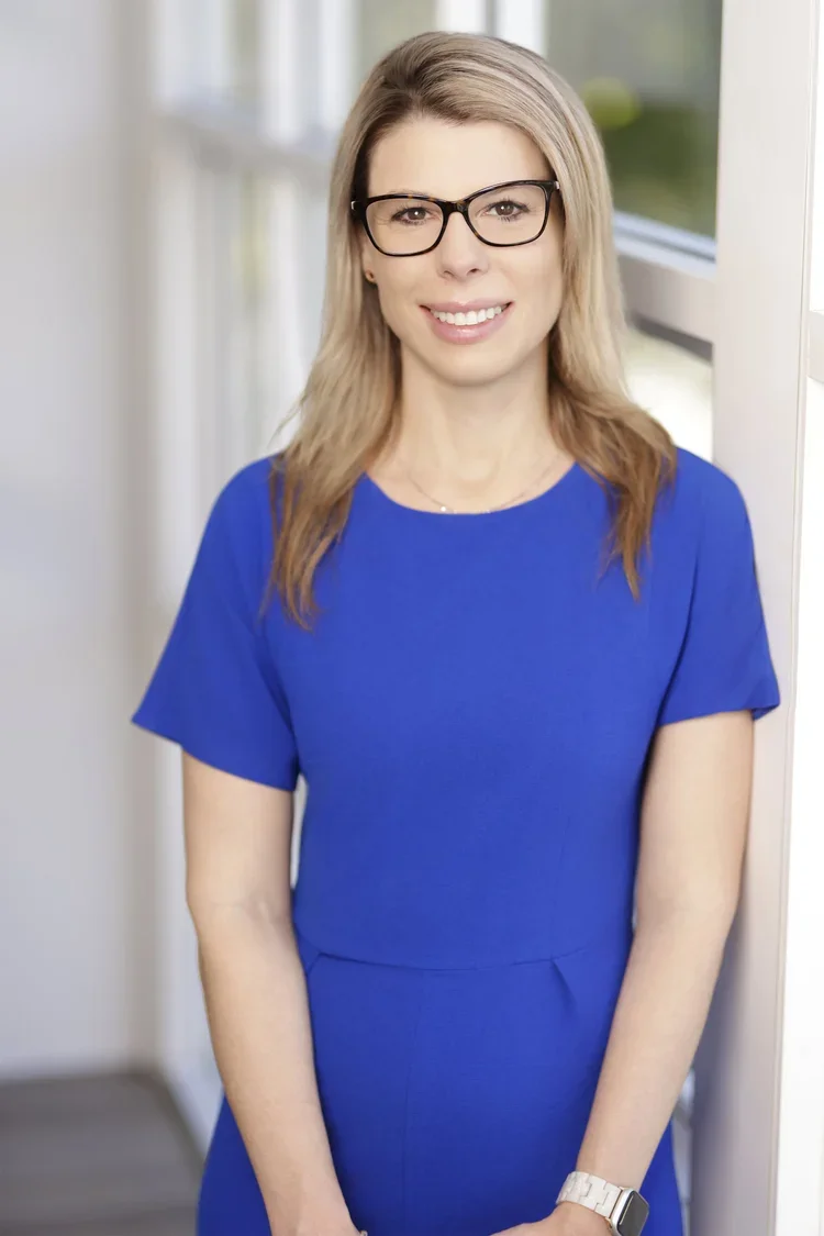 A woman with blonde hair wearing glasses and a blue dress, standing indoors near a window, smiling at the camera.