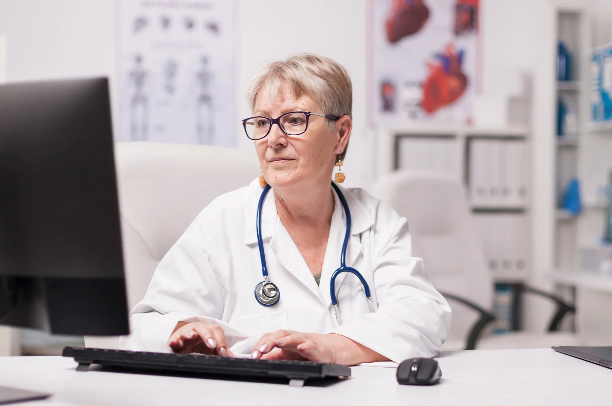 A female doctor with short hair, glasses, and a stethoscope around her neck is sitting at a desk working on a computer in a medical office or clinic, with medical posters and shelves in the background.
