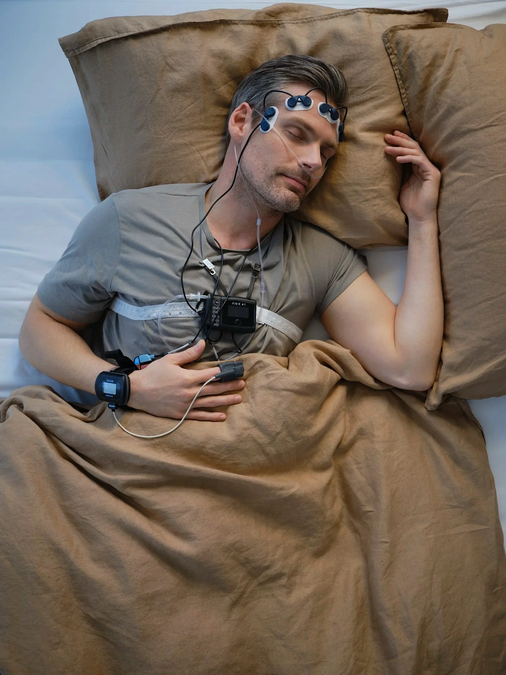 A man is sleeping on a bed with his head resting on a large pillow. He is wearing a gray t-shirt and a medical device with wires attached to his head and chest, indicating he is undergoing a sleep study or medical monitoring.