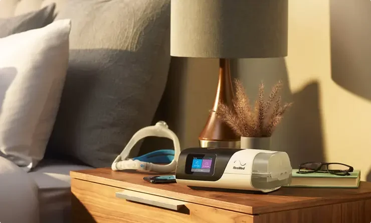 Nightstand with a CPAP (Continuous Positive Airway Pressure) device, glasses, a book, a smartphone, a lamp, and decorative plant in a bedroom.
