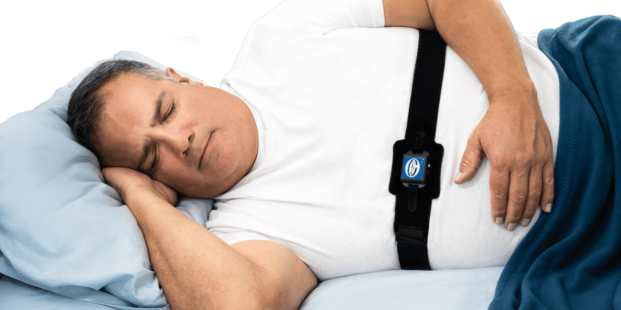 A man sleeping on his side in bed, wearing a white t-shirt and a sleep monitor on his chest.