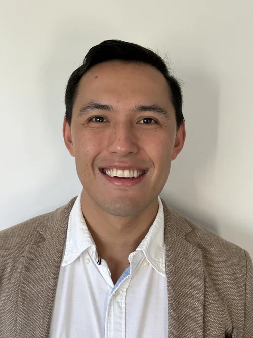 A smiling man with short dark hair wearing a beige blazer and white shirt against a plain white background.