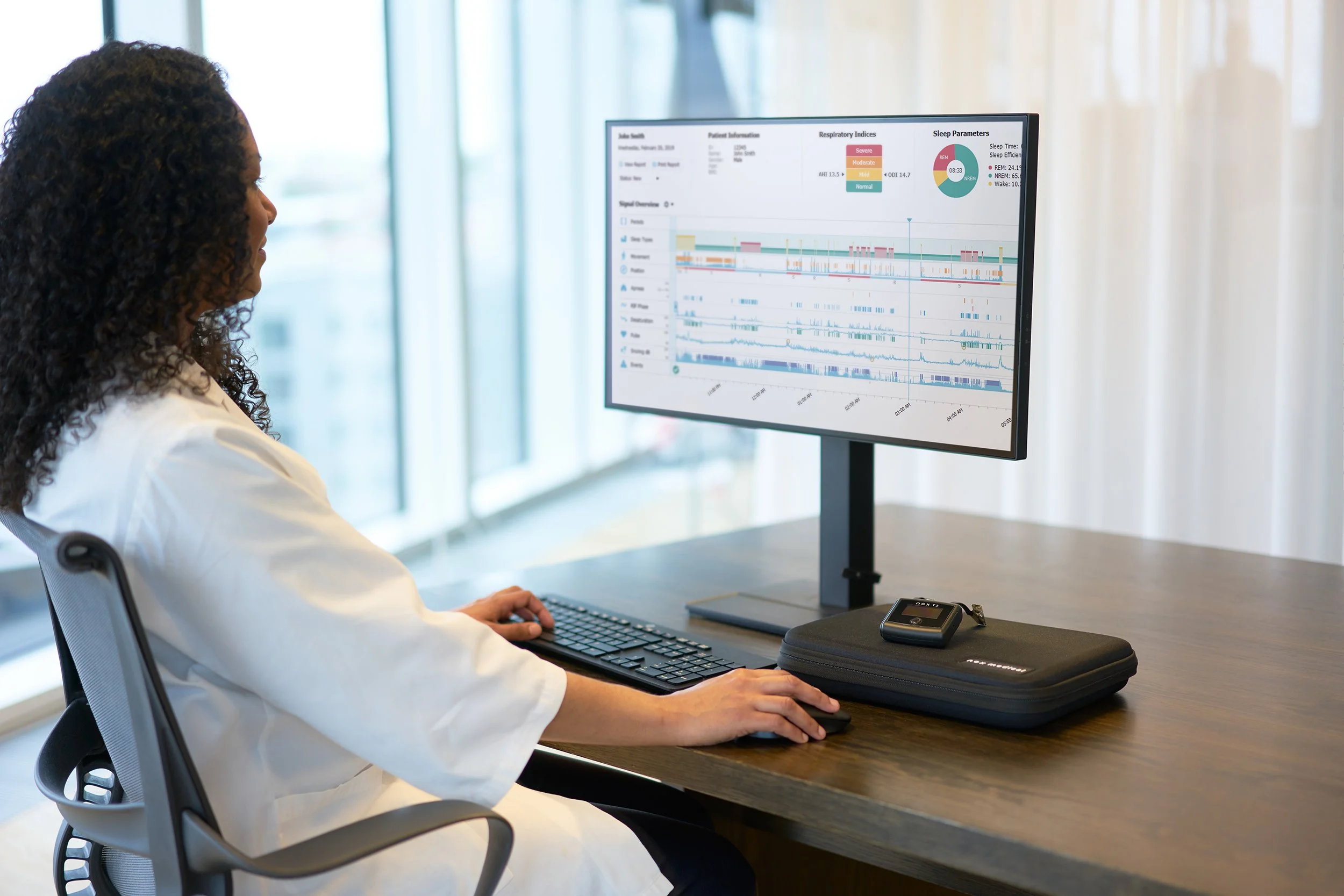 A woman with curly hair in a white coat sitting at a desk looking at a computer monitor displaying a patient health analysis chart.