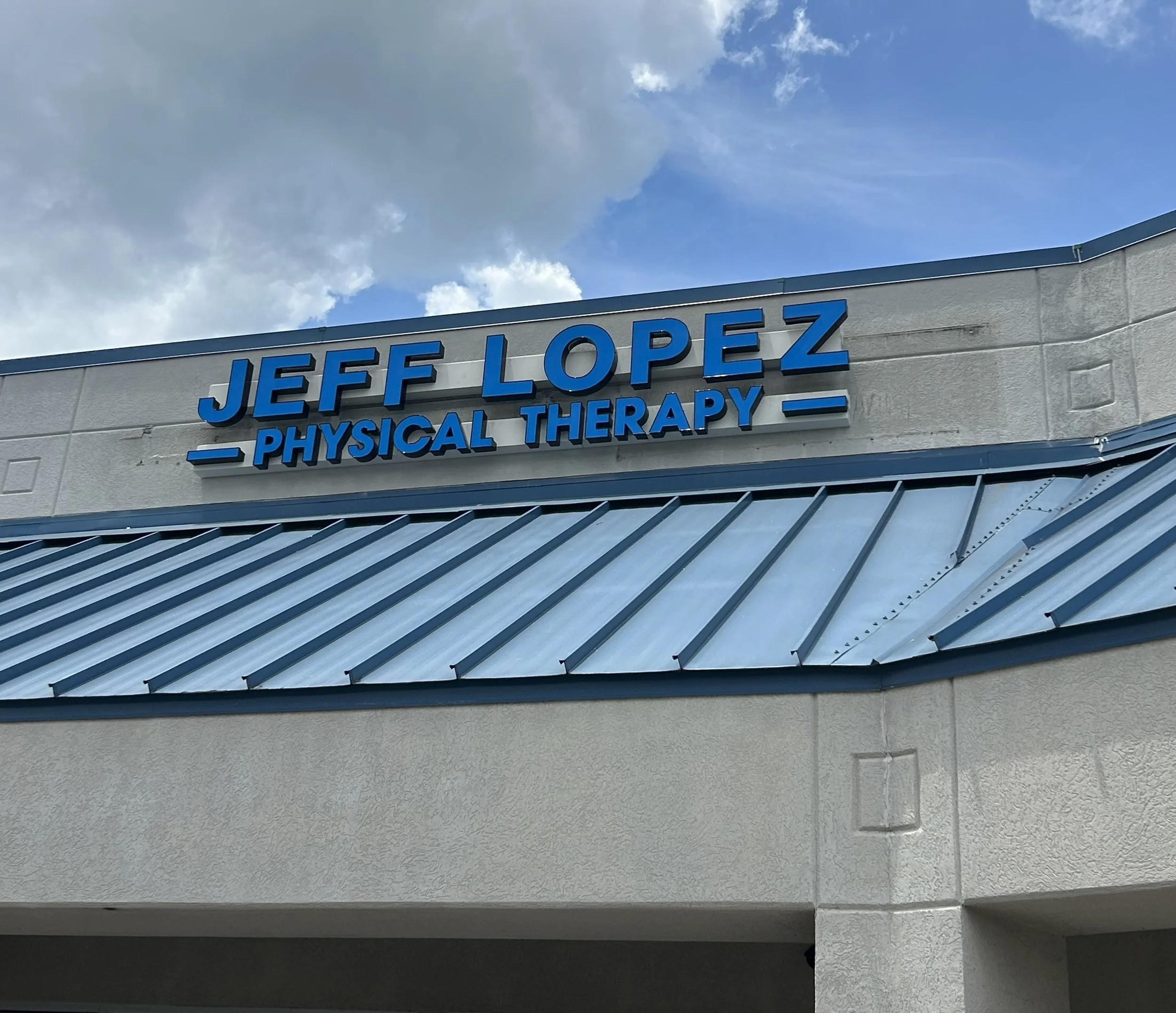 Sign on building that reads 'Jeff Lopez Physical Therapy' with a blue sky and clouds in the background.