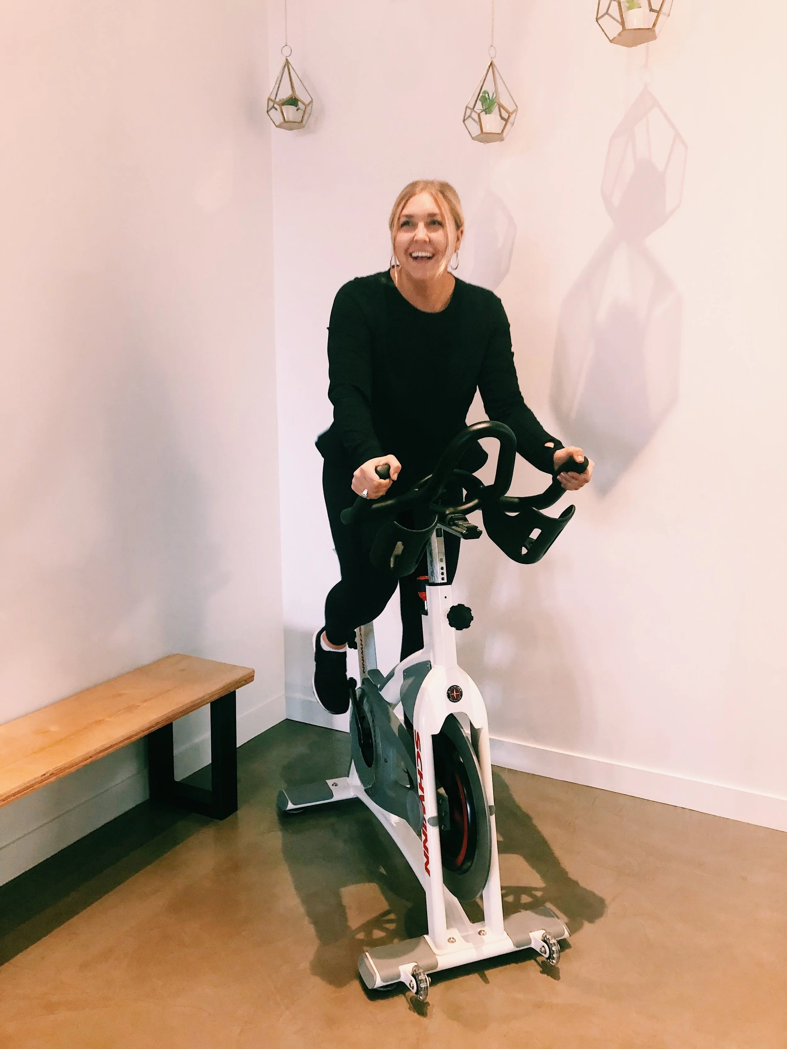 A woman riding an exercise bike indoors, smiling, with a white wall and hanging geometric planters with small plants behind her.