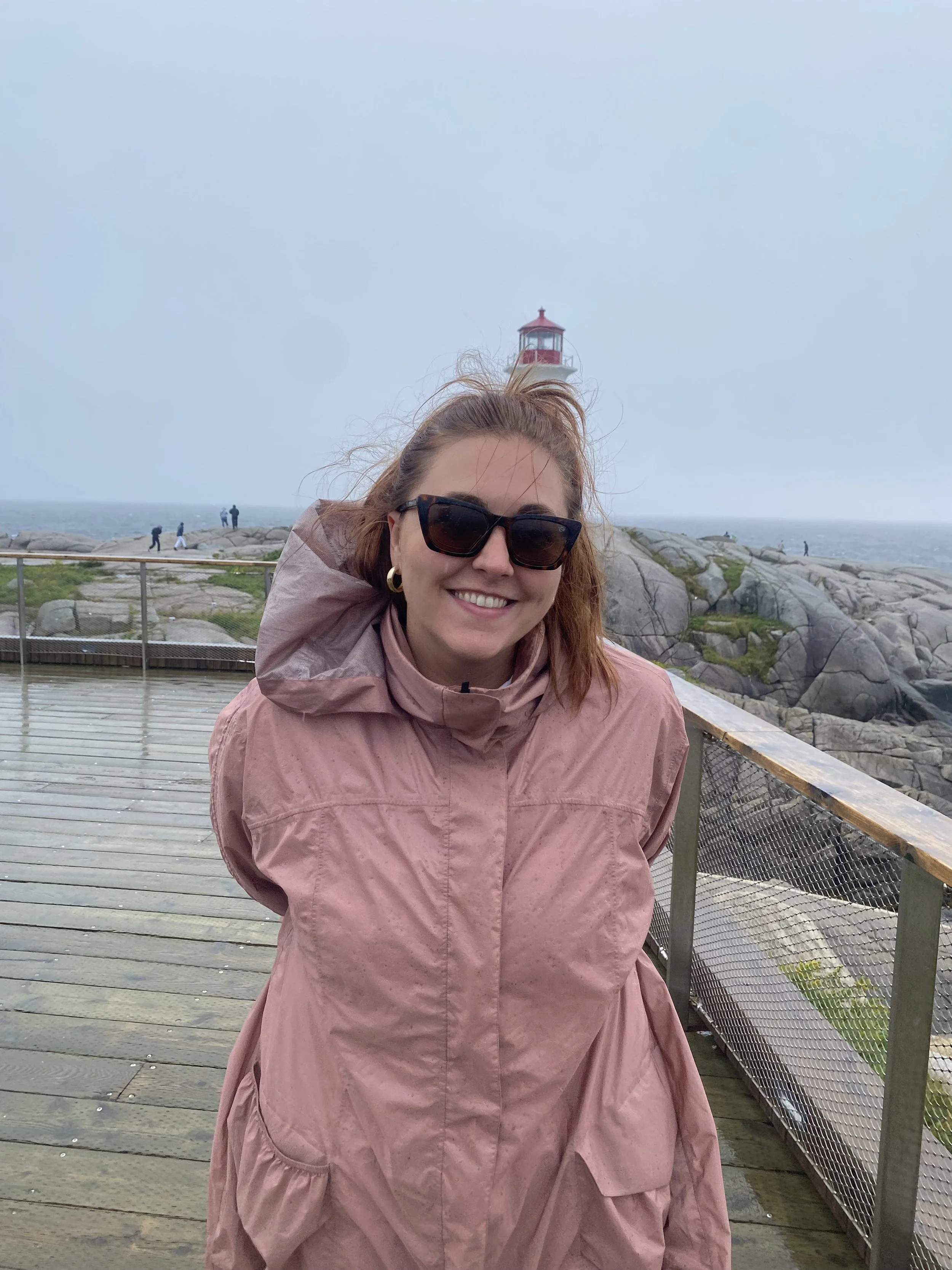 A woman with red hair, wearing sunglasses and a pink jacket, smiling on a wooden boardwalk near the water with a lighthouse in the background.