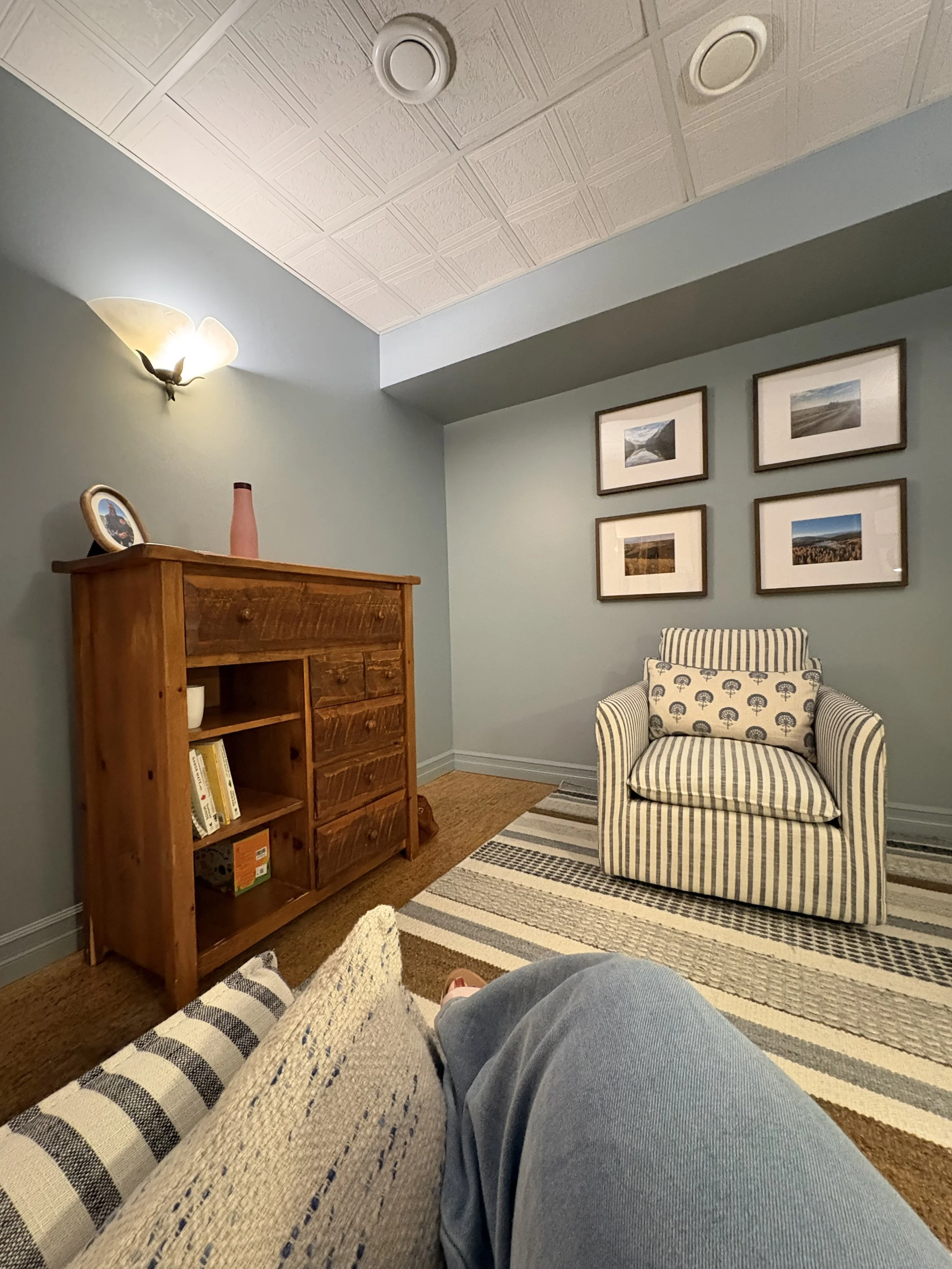 The image shows a cozy living room with a person sitting on a chair, wearing blue jeans and brown shoes, with their legs extended over a striped woven table runner on a plaid fabric ottoman. In the background, there's a wooden bookshelf, a framed photograph, a pink vase, a wall-mounted lamp, and four landscape photographs on the wall. The ceiling is white with decorative tiles, and the room has a hardwood floor with a striped area rug.
