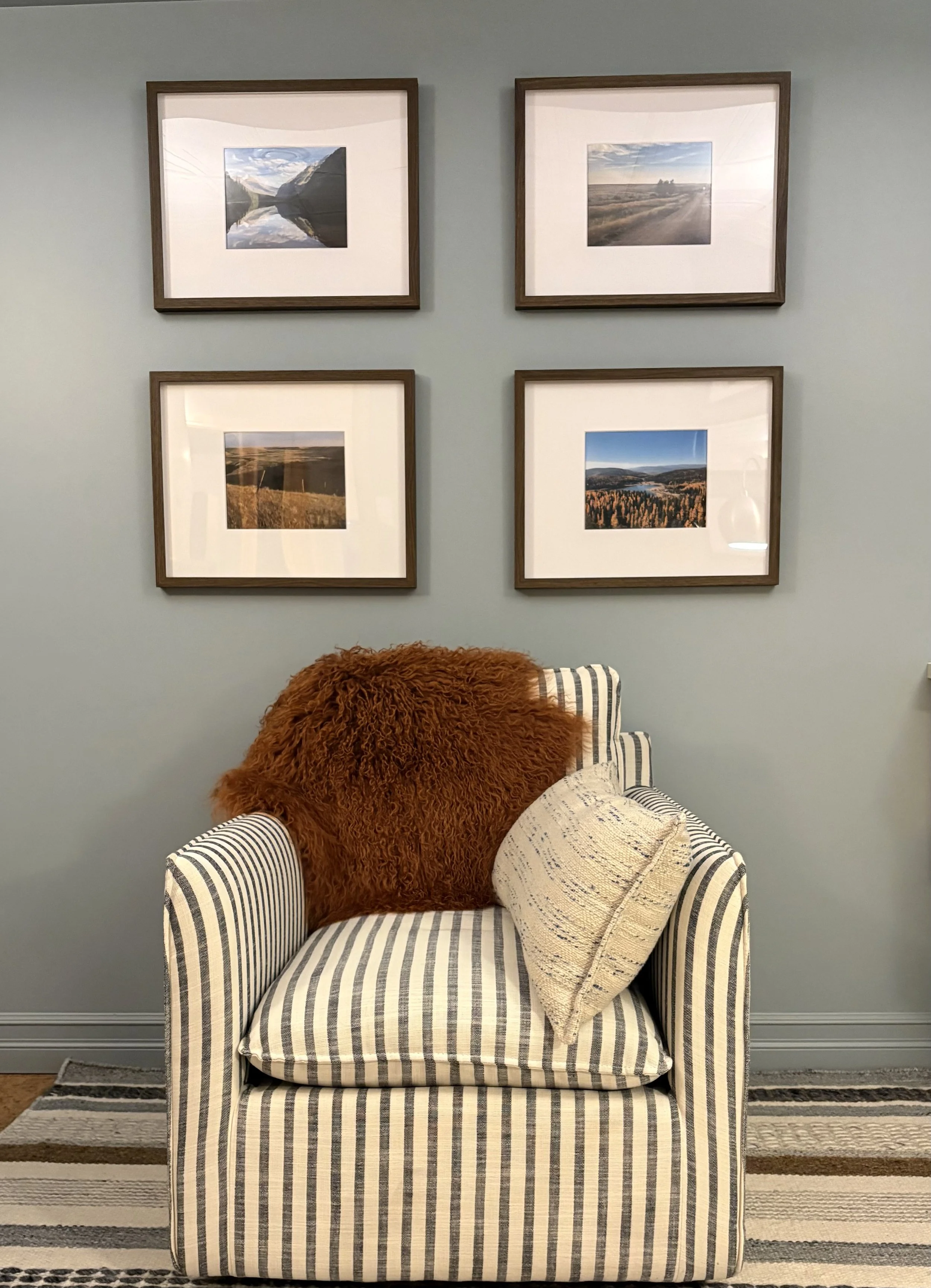Living room scene with a striped armchair, a beige pillow, a brown furry throw, a striped rug, a blue wall, and four framed landscape photographs.